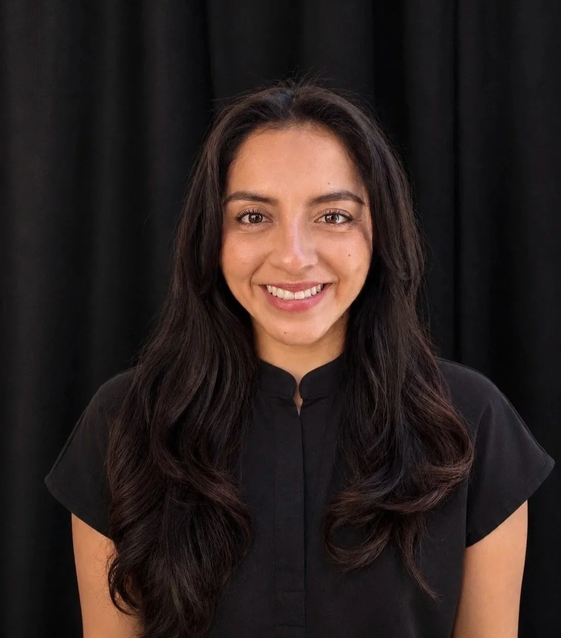 A young woman with long, wavy dark hair smiling at the camera, wearing a black top against a black curtain background.