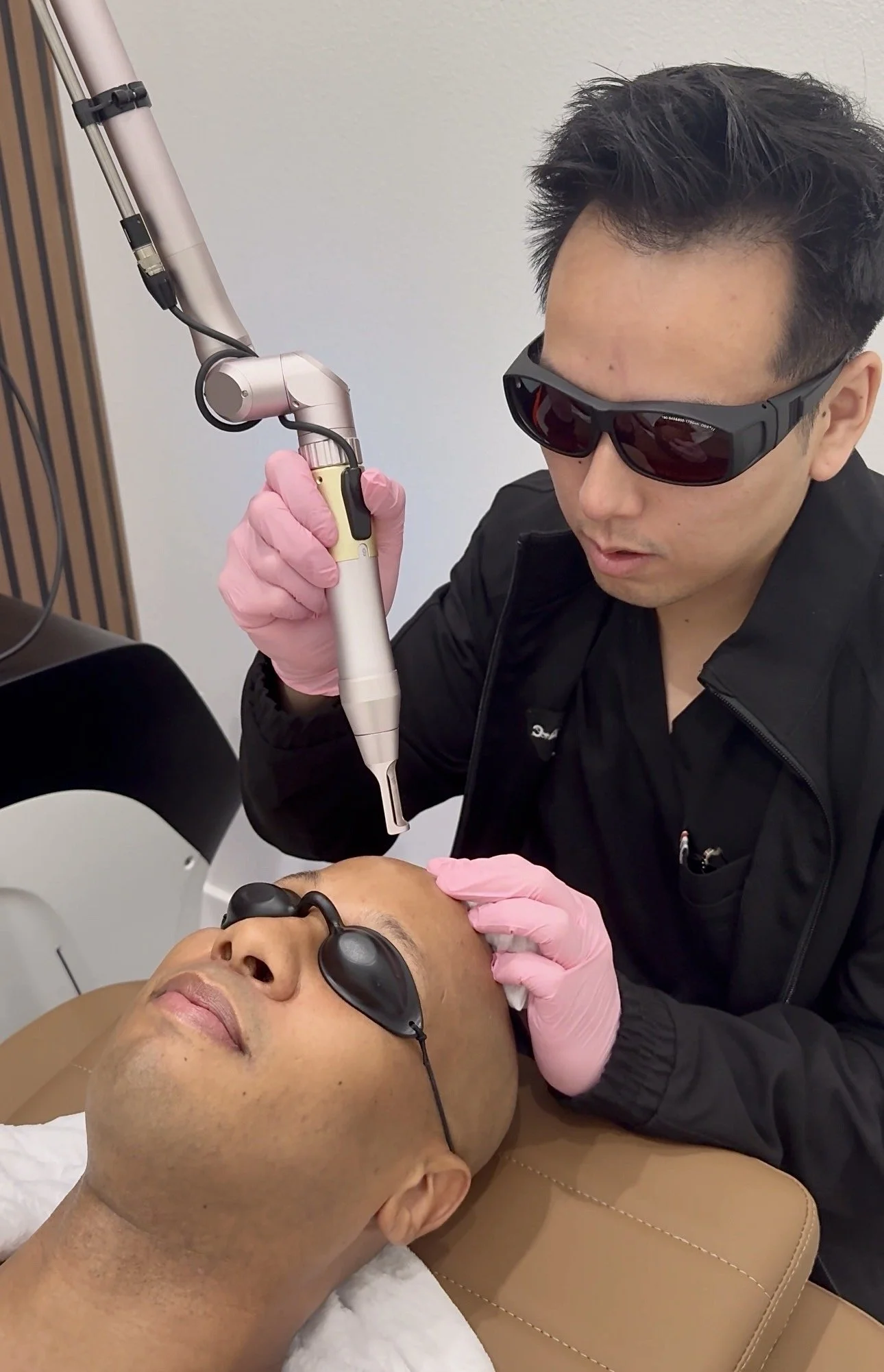 A man lying down with protective glasses, receiving a cosmetic or dermatological treatment from a practitioner holding a device near his scalp, in a clinical setting.