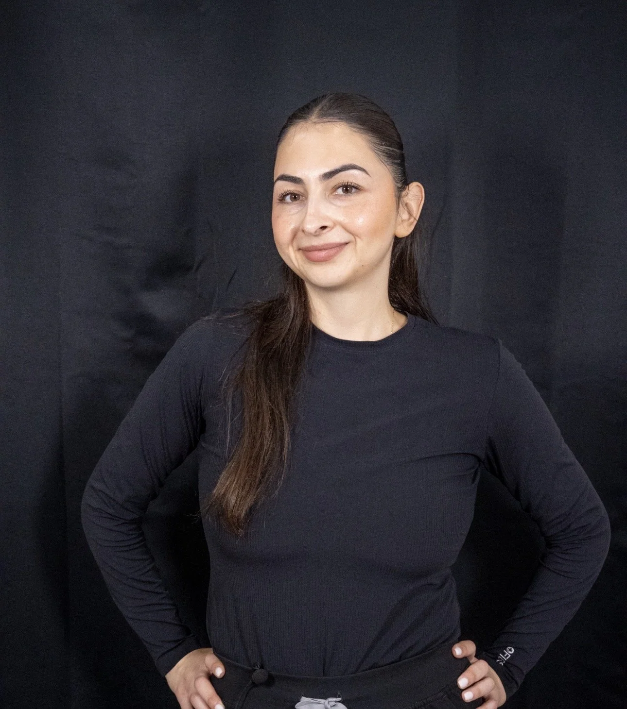 A woman with long brown hair wearing a black long-sleeve shirt standing against a black backdrop, with her right hand on her hip and smiling slightly.
