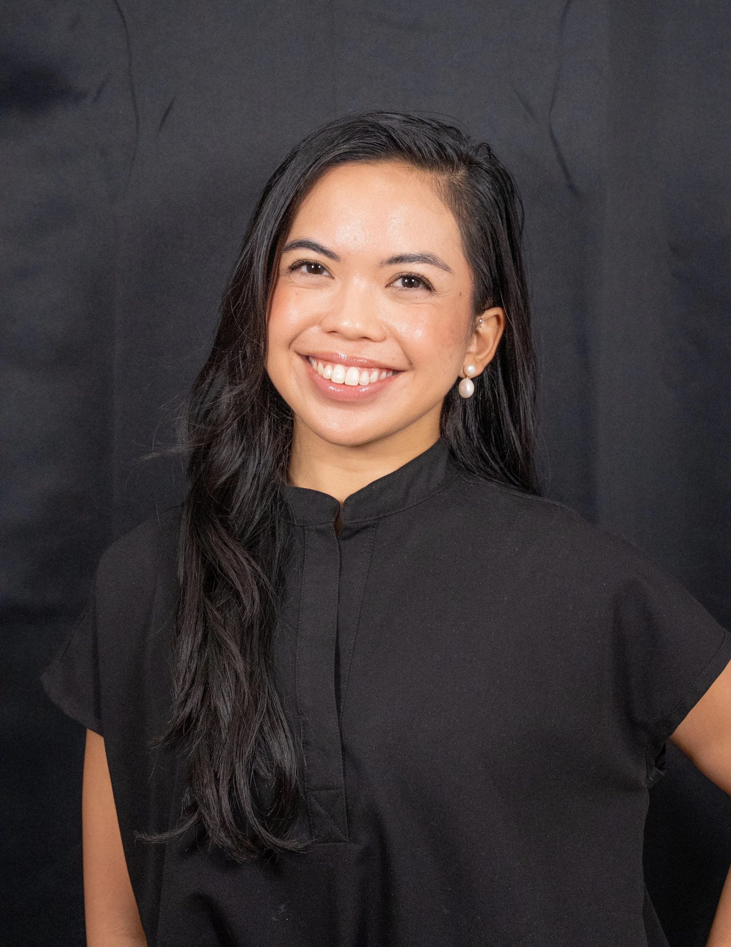Close-up of a woman with long dark hair, wearing a black outfit and pearl earrings, smiling against a dark background.
