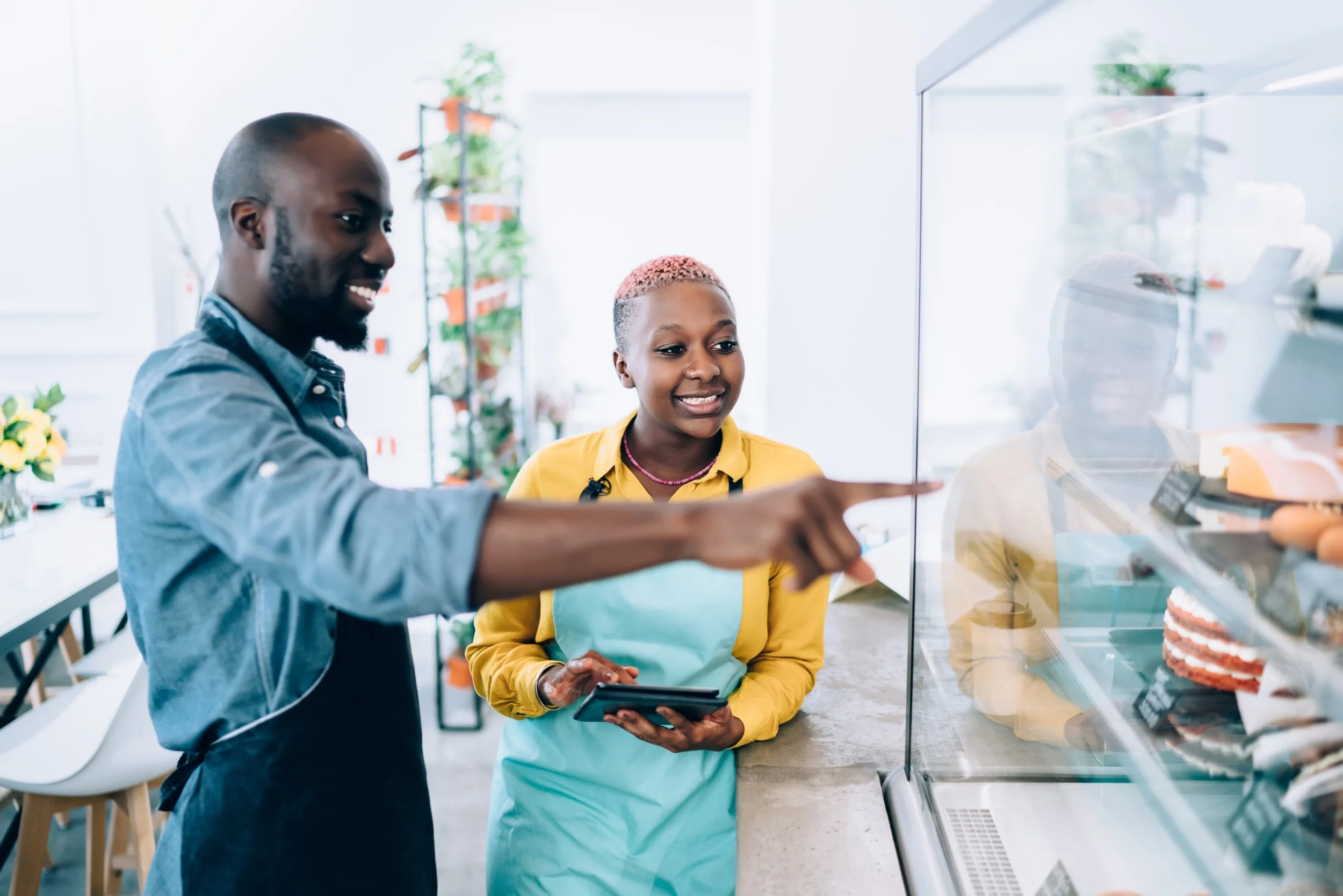 Two people, a man and a woman, looking at cakes in a bakery display case. The man is pointing at the cakes and the woman is holding a phone, both smiling.