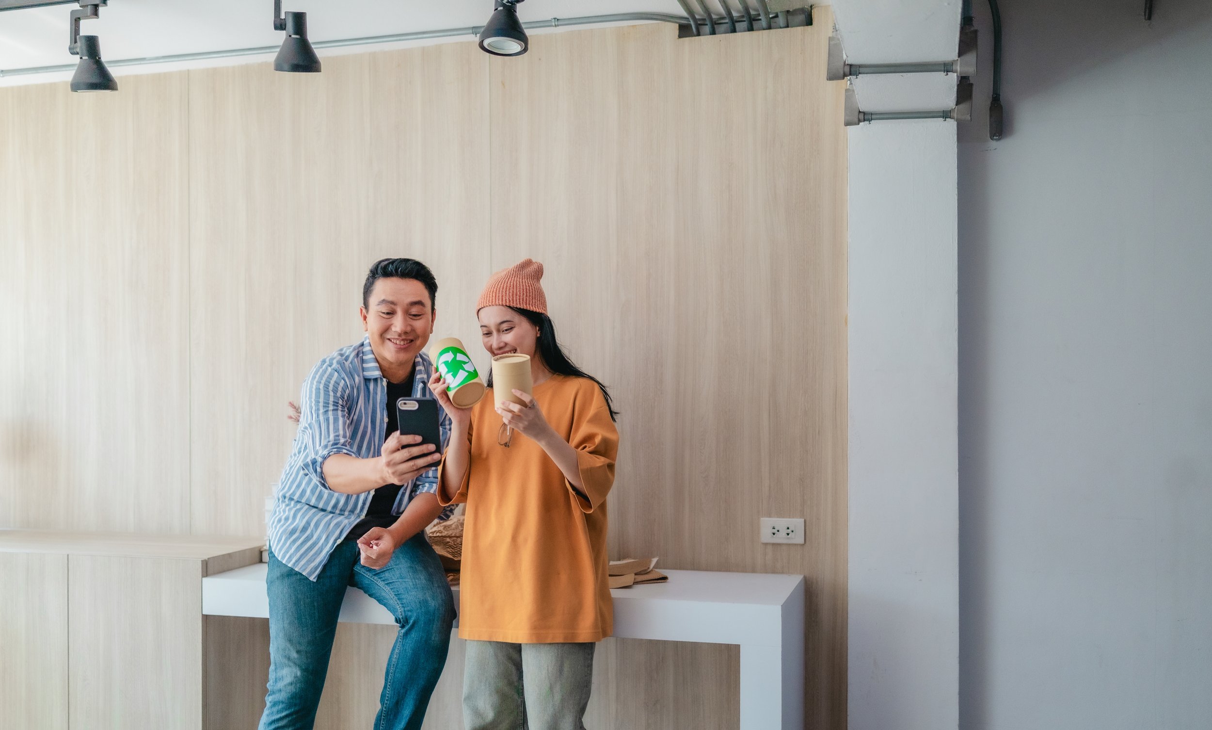 Two people, a man and a woman, are standing indoors in front of a light wood-paneled wall with black ceiling lights above. They are smiling and looking at a smartphone the man is holding. Both are holding disposable coffee cups.