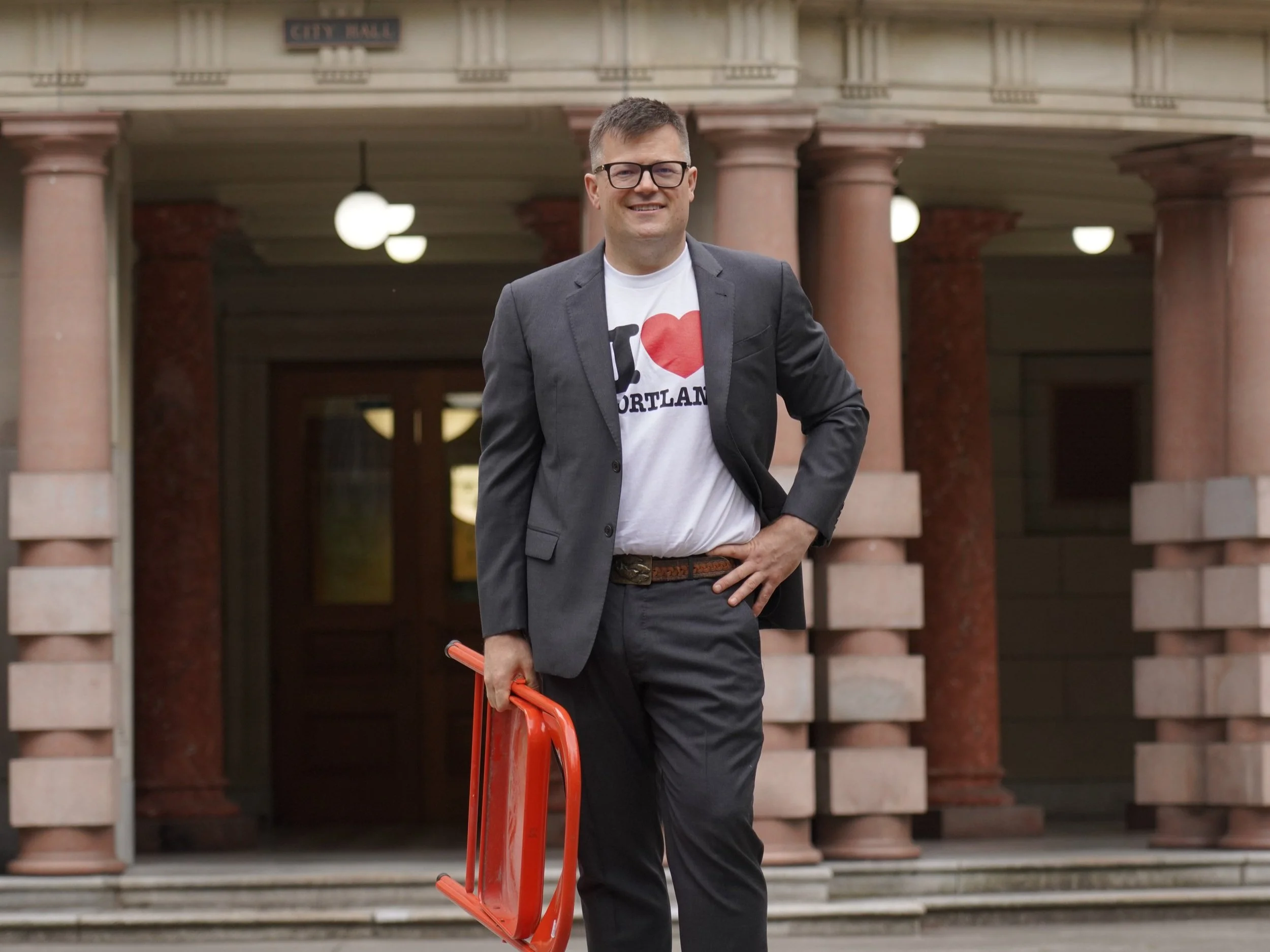Photo of Josh Leake, Candidate, Mayor of Portland in front of Portland City Hall with an orange folding chair.