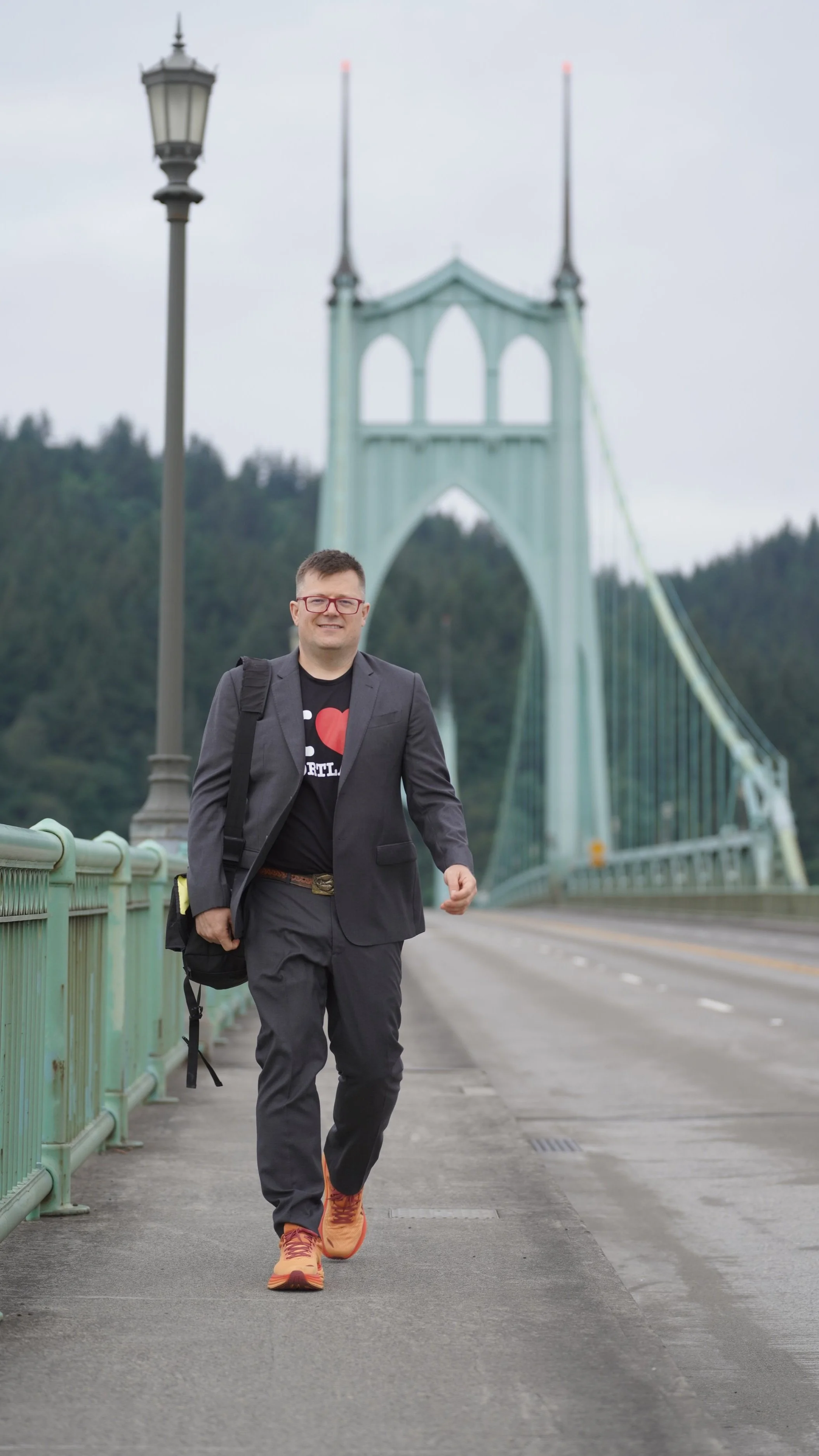 A man walking on a bridge with a green rail, wearing a dark suit, black t-shirt, orange shoes, and carrying a black backpack, with a large bridge structure and cloudy sky in the background.