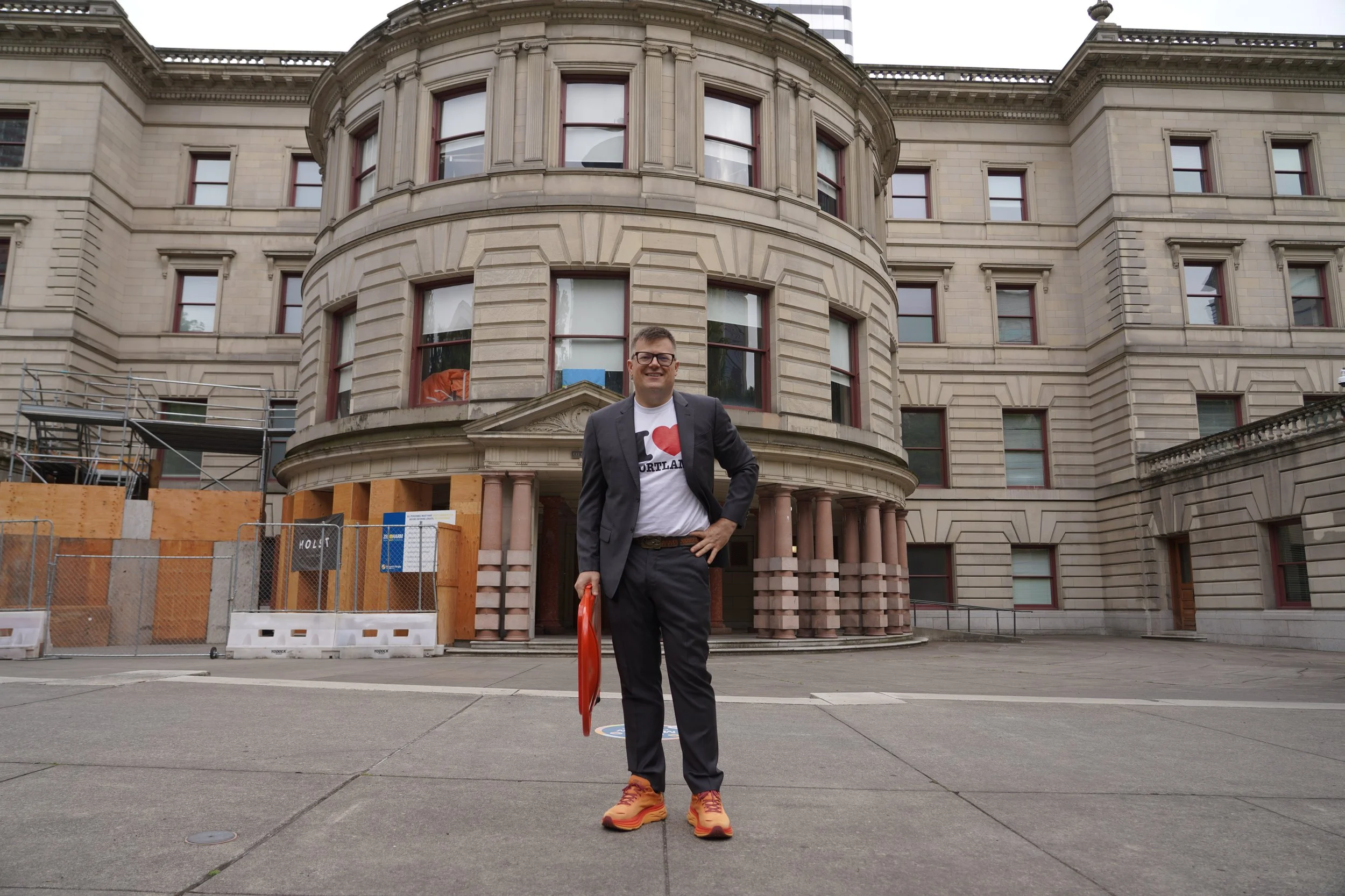 A man in a suit and glasses smiling, standing outdoors in front of a historic stone building with multiple windows, wearing a white T-shirt with a red heart and the word 'Portland', orange shoes, and holding an orange object.