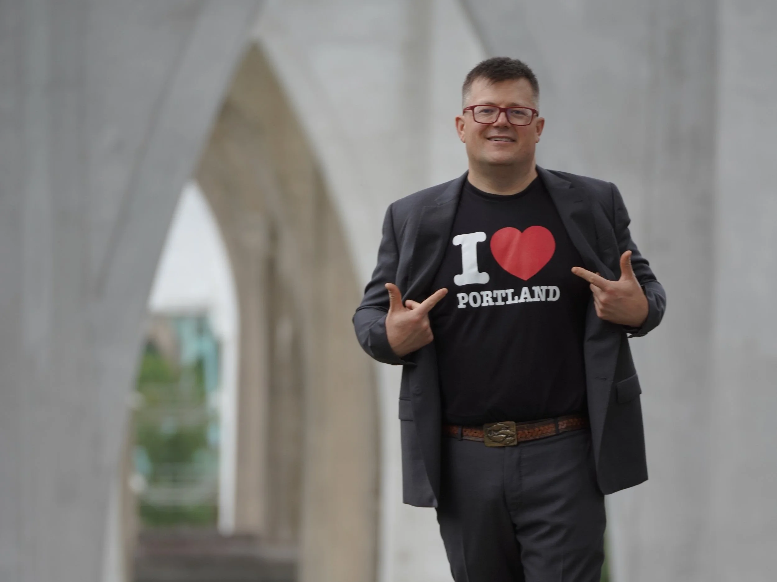 A man smiling and pointing at his black t-shirt with the words "I ❤️ PORTLAND" in white and red text. He is wearing glasses, a blazer, and a belt. The background shows an architectural structure with a blurred walkway and a cloudy sky.