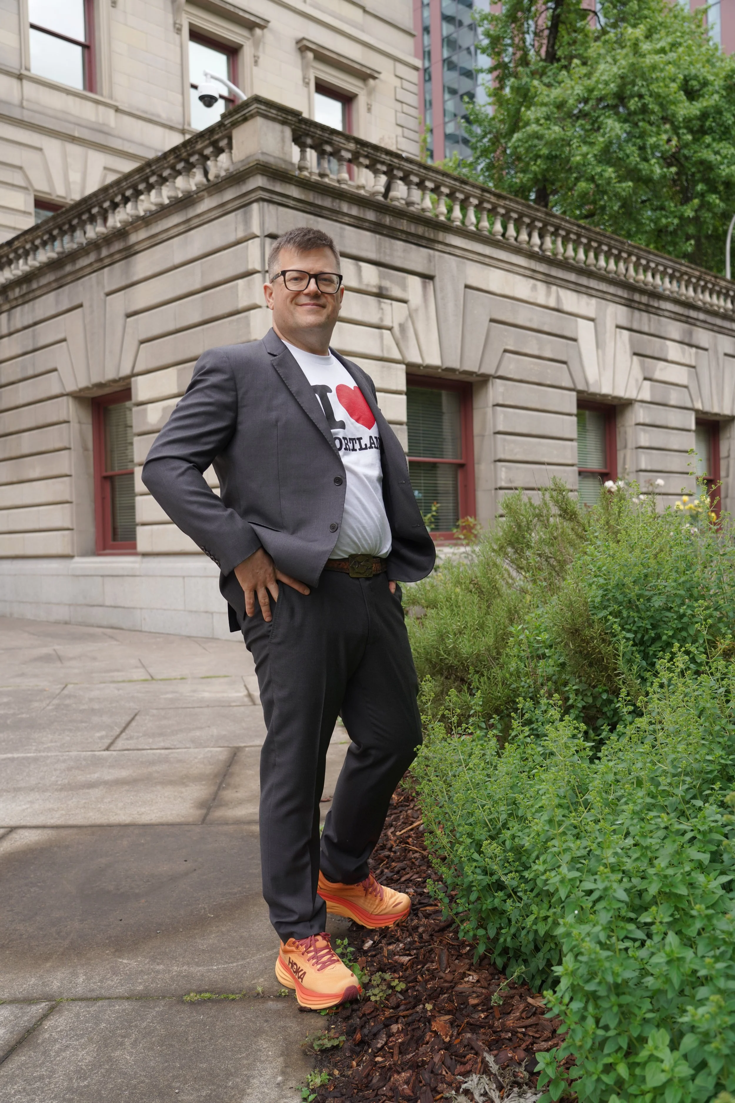 A man in a suit and glasses standing outdoors on a city sidewalk, with a historic stone building, trees, and greenery in the background. He is wearing a graphic T-shirt, dress pants, and colorful sneakers.