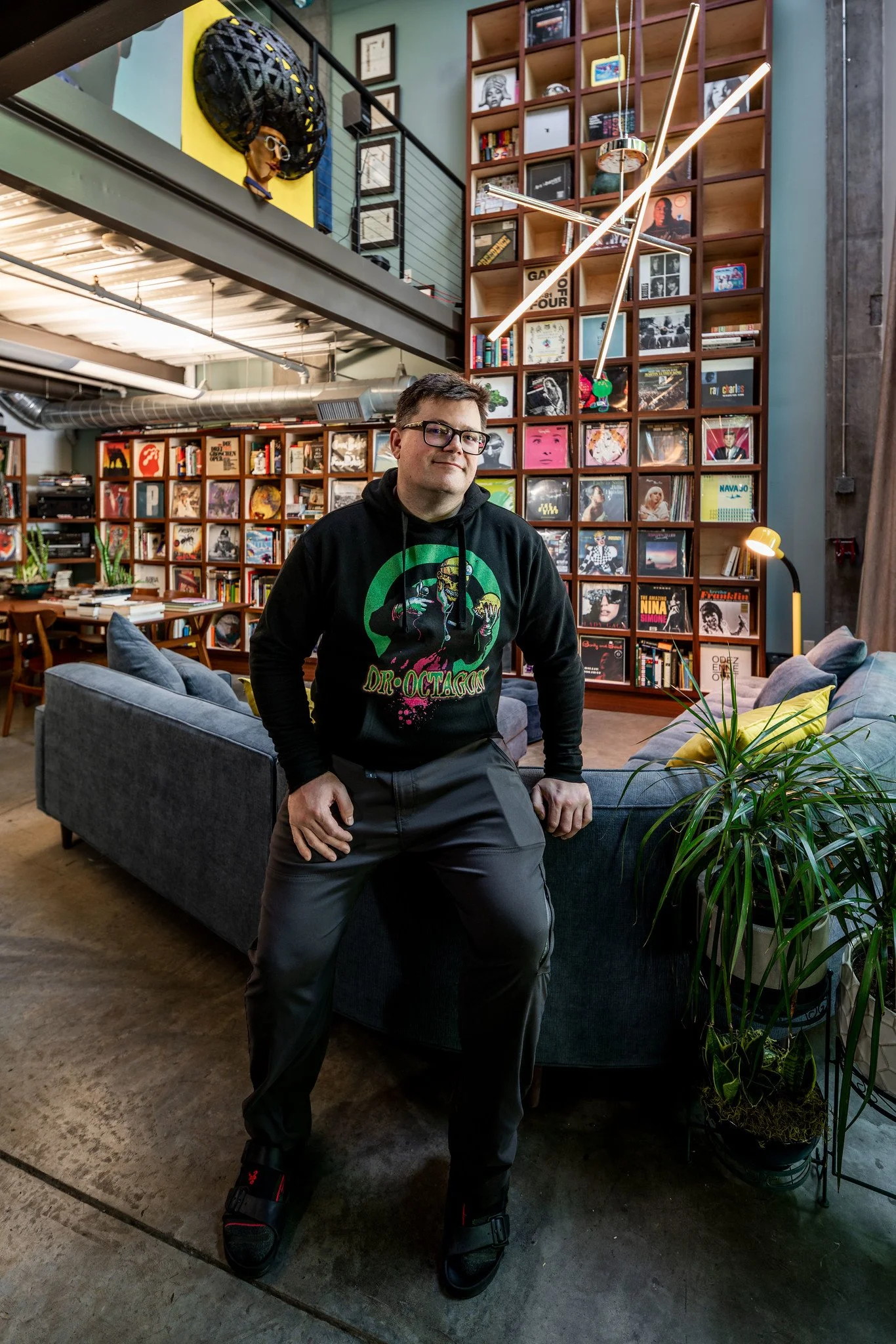 A man wearing glasses, a black hoodie featuring a colorful graphic of a skeleton, and black pants, seated on a dark sofa in a stylish, modern room with wooden shelves filled with books and vinyl records, a decorative yellow cushion, houseplants, and contemporary lighting fixtures.