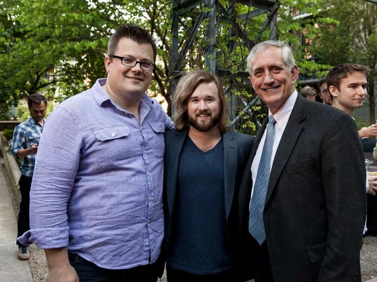 Three men standing together outdoors at a social event, smiling for the camera. Two in casual shirts and one in a suit, with a background of greenery and other people.