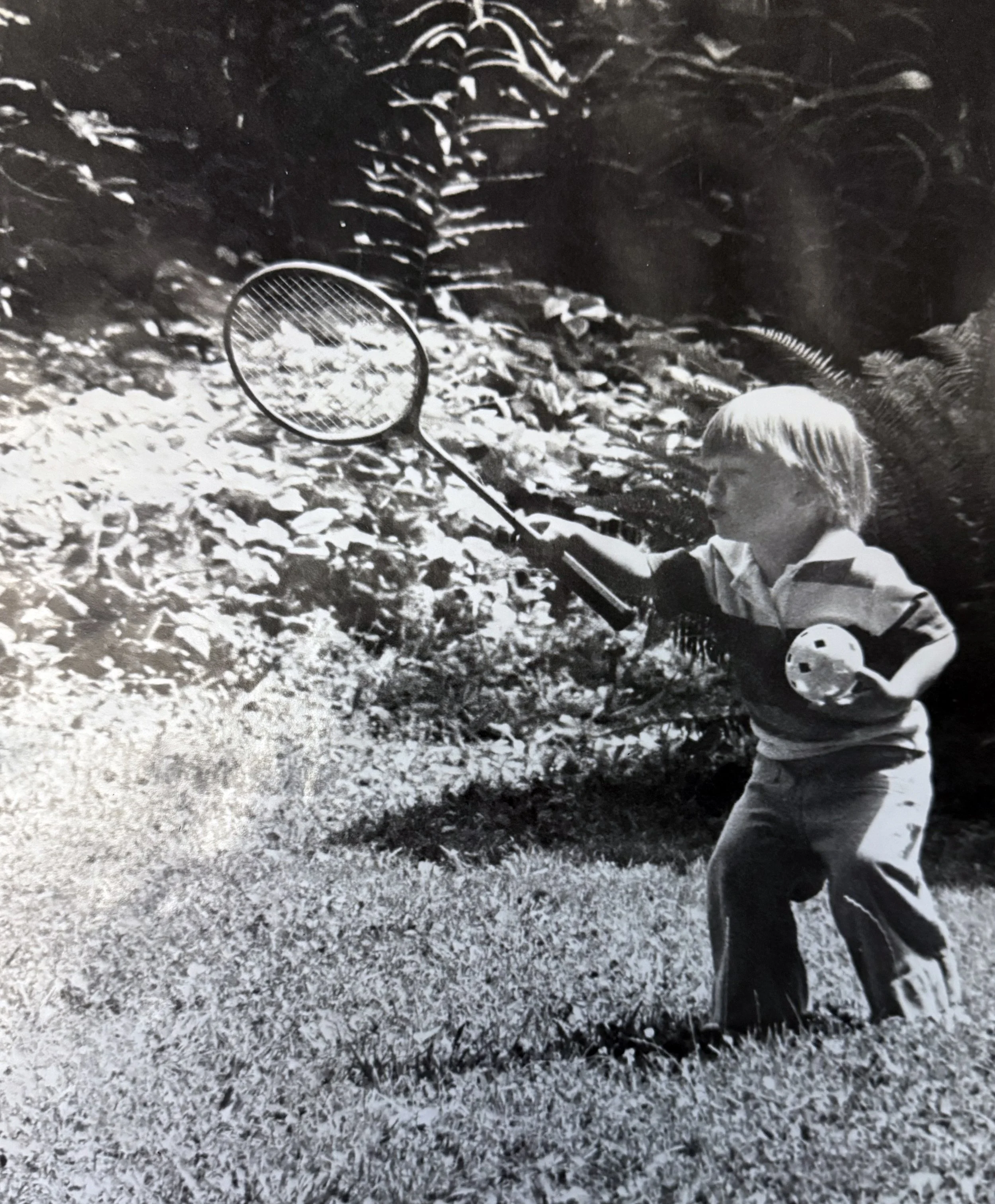 A young boy with blonde hair, wearing a long-sleeved shirt and pants, holding a butterfly net, standing on grass with small rocks, outdoors near dense bushes or trees.