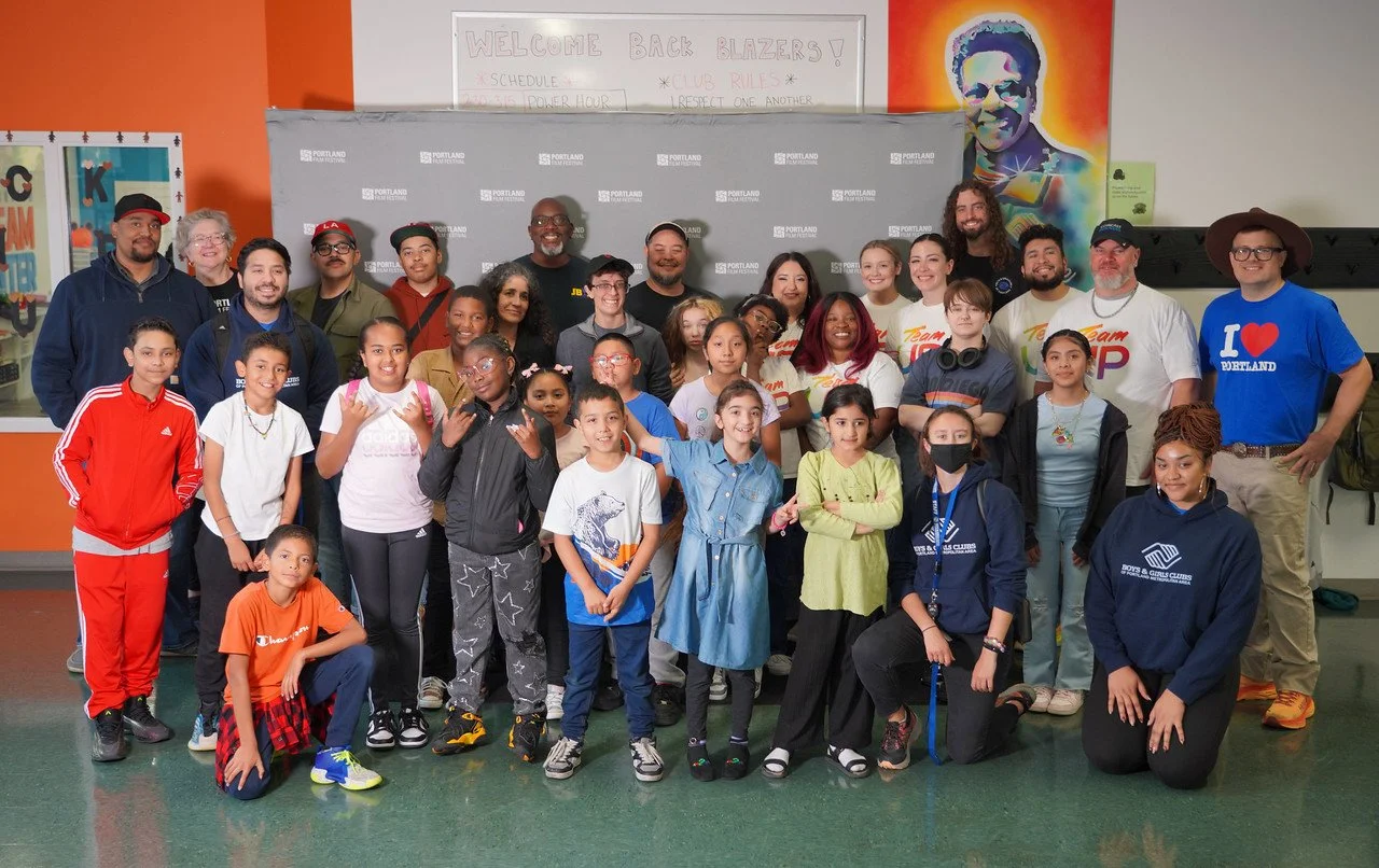 Group photo of children and adults in a school hallway, with a gray backdrop, a colorful mural of a person, and a whiteboard with writing behind them.