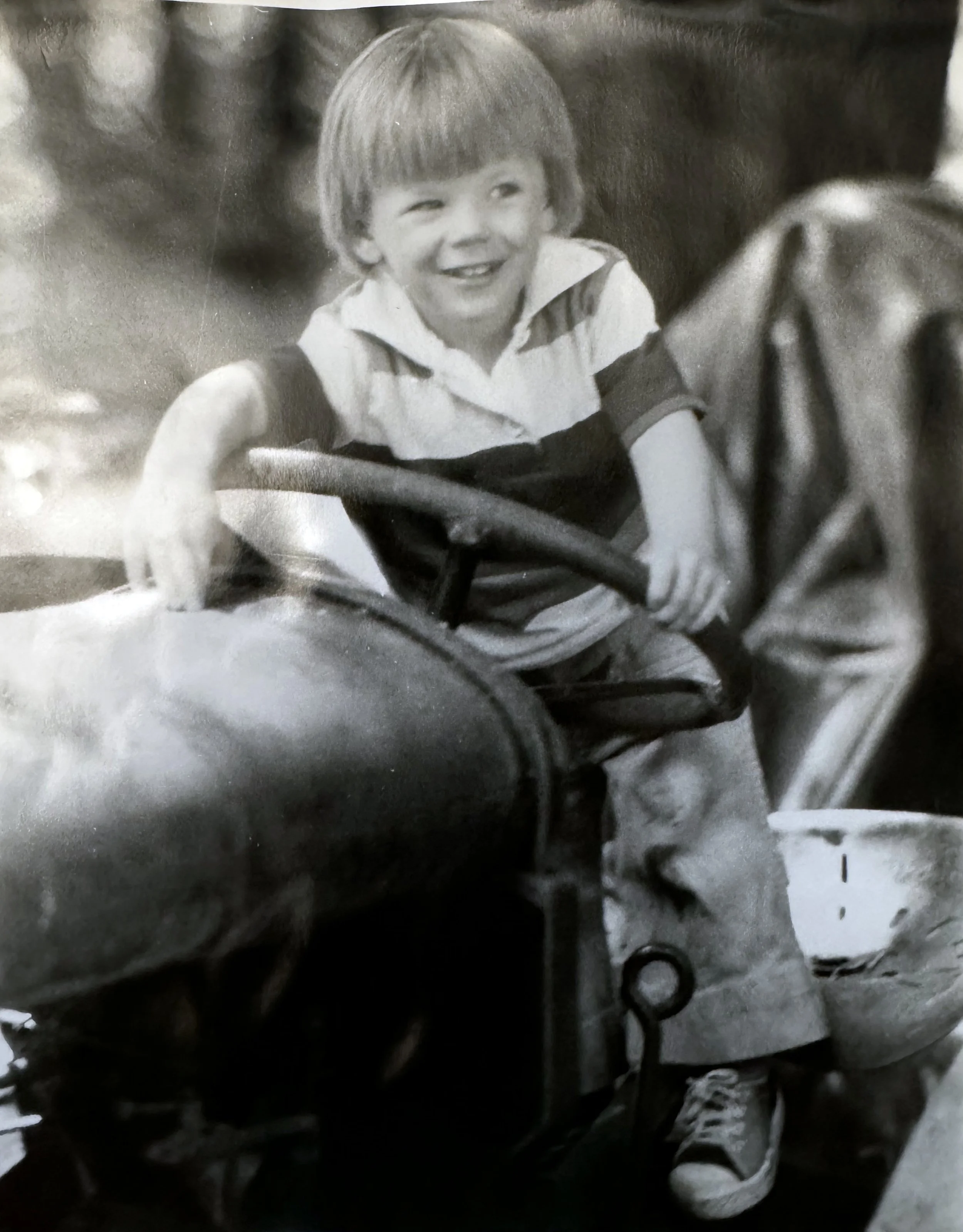 A young boy with a short haircut smiling and reaching out with his right arm. He is wearing a white and dark striped polo shirt and sneakers. The background appears to be an indoor space with some equipment or objects.