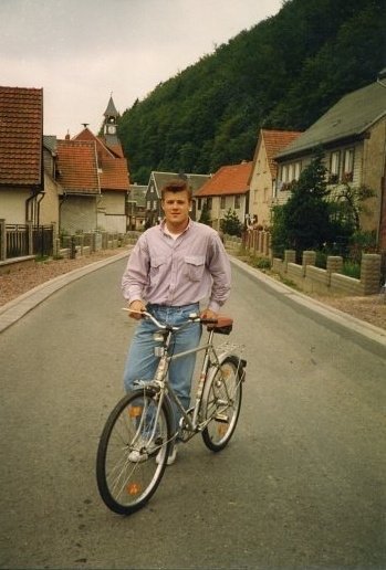 A young man standing with a bicycle on a quiet street in a small town, with houses and a clock tower in the background, and a green hill on the right side.