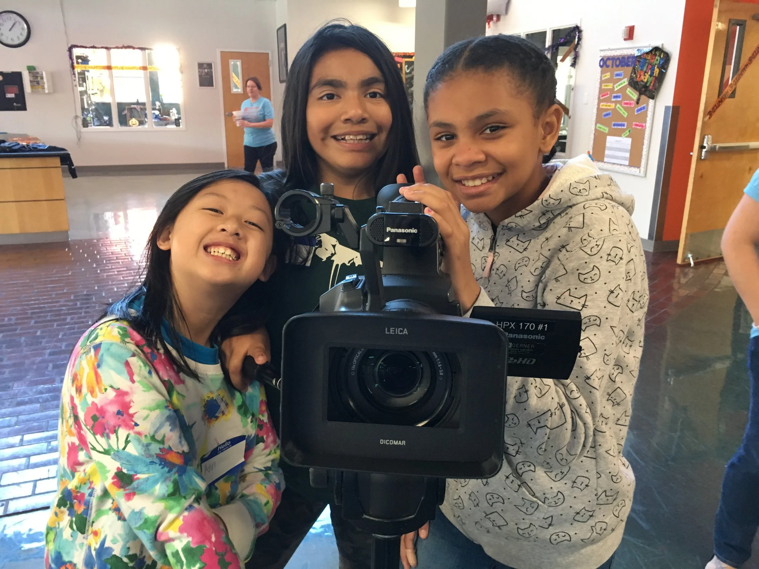 Three children, two girls and one boy, smiling and gathered around a video camera in a classroom or community center with bulletin boards, a clock, and a woman in the background.