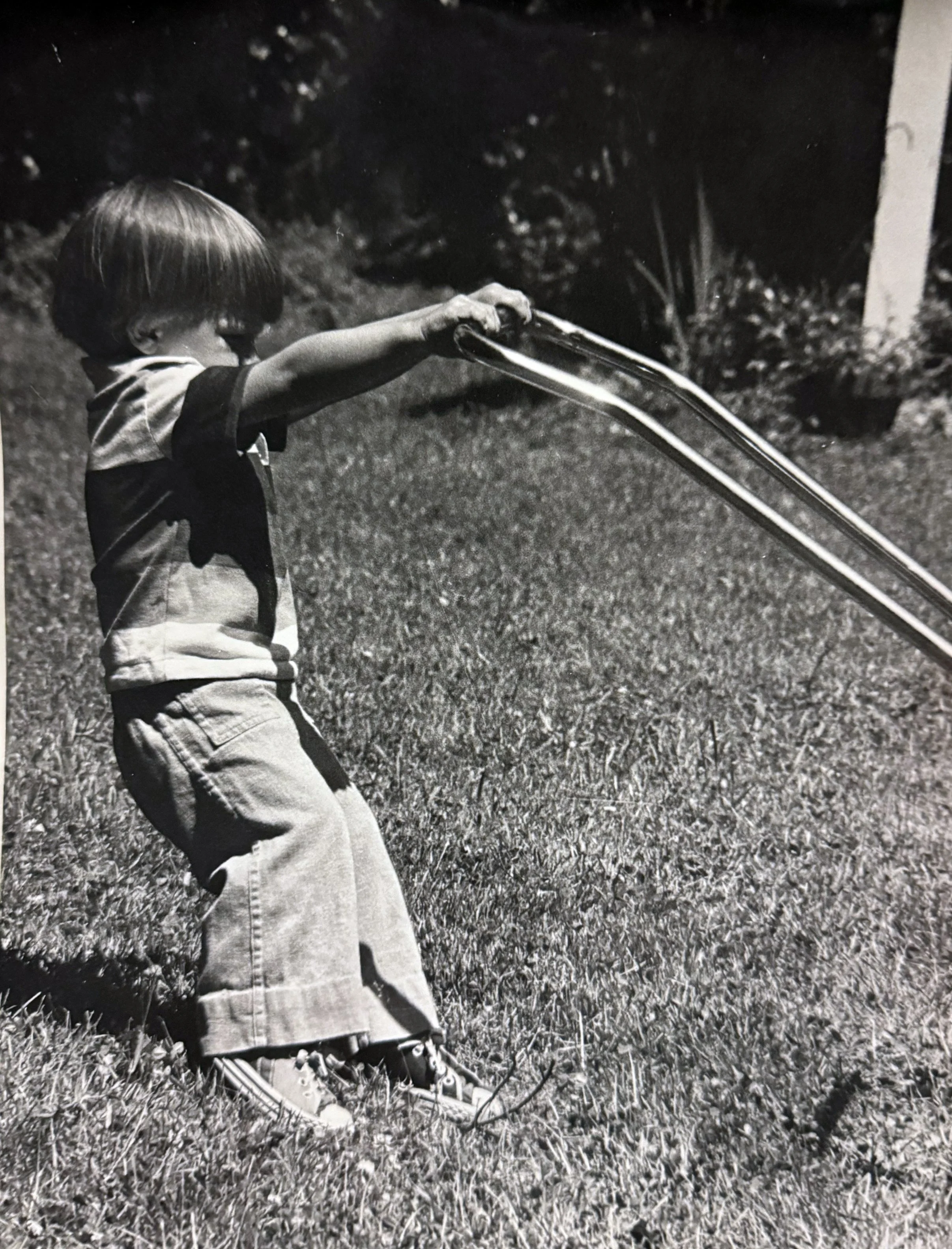 A young boy with a short haircut, wearing a t-shirt, shorts, and sneakers, is outdoors in a grassy area, reaching out to touch or hold a pair of metal rails in a playful manner.