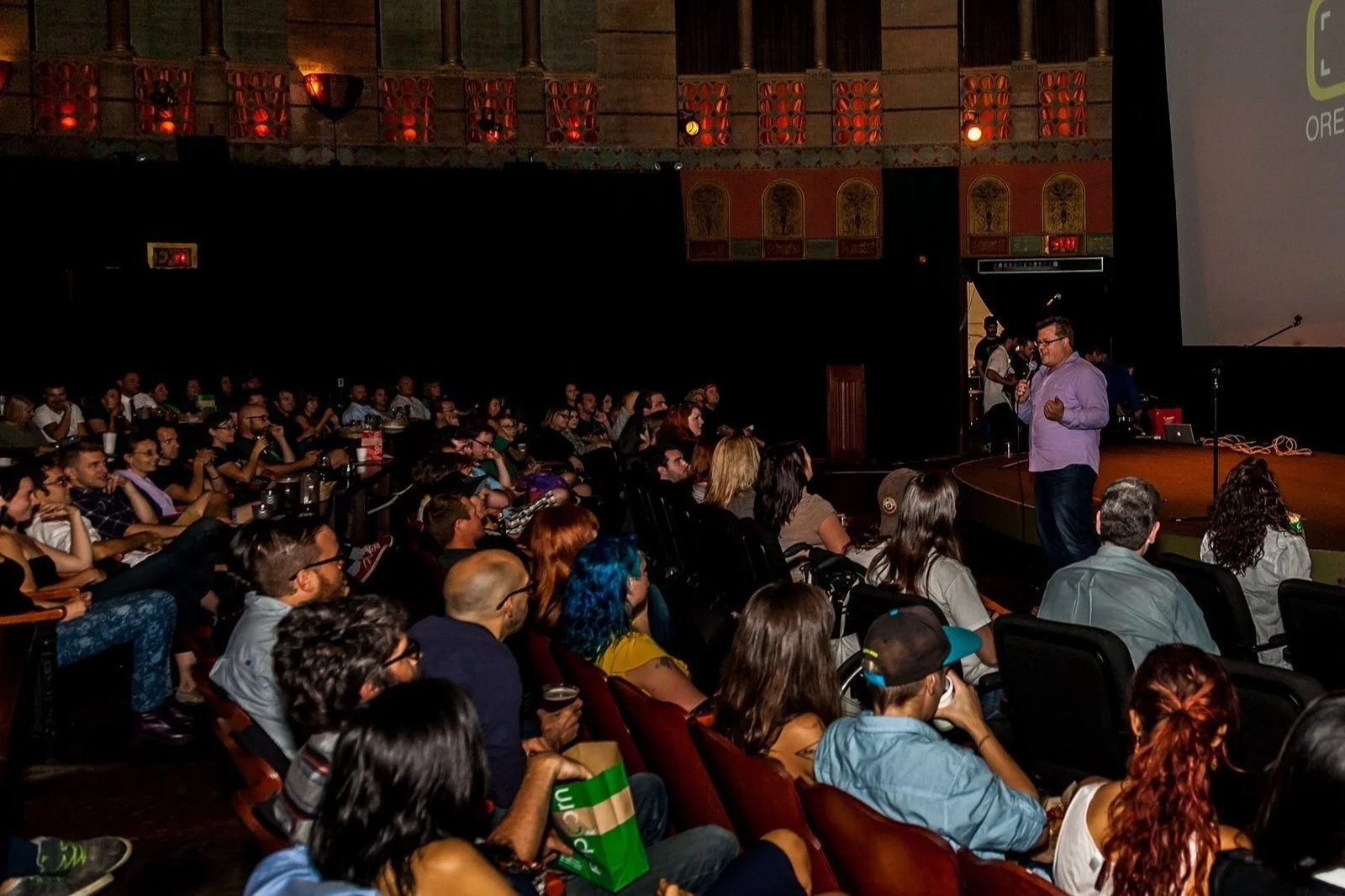 A man in a purple shirt holding a microphone is speaking on stage in front of a large audience sitting in a theater or auditorium. The audience appears attentive, and the setting is dimly lit with a large projection screen behind the speaker.