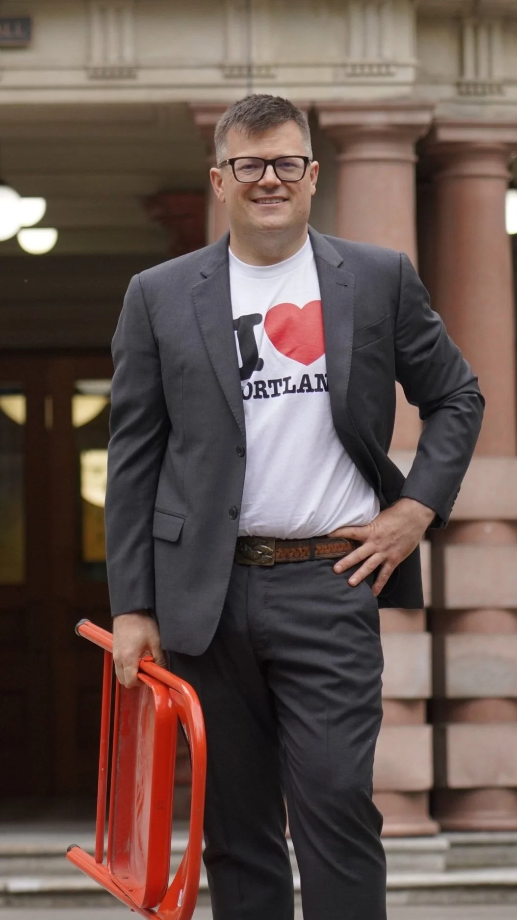 A man smiling, wearing glasses, a gray suit jacket, white T-shirt with a red heart and black text, gray pants, holding an orange traffic cone, standing in front of a building with large columns.