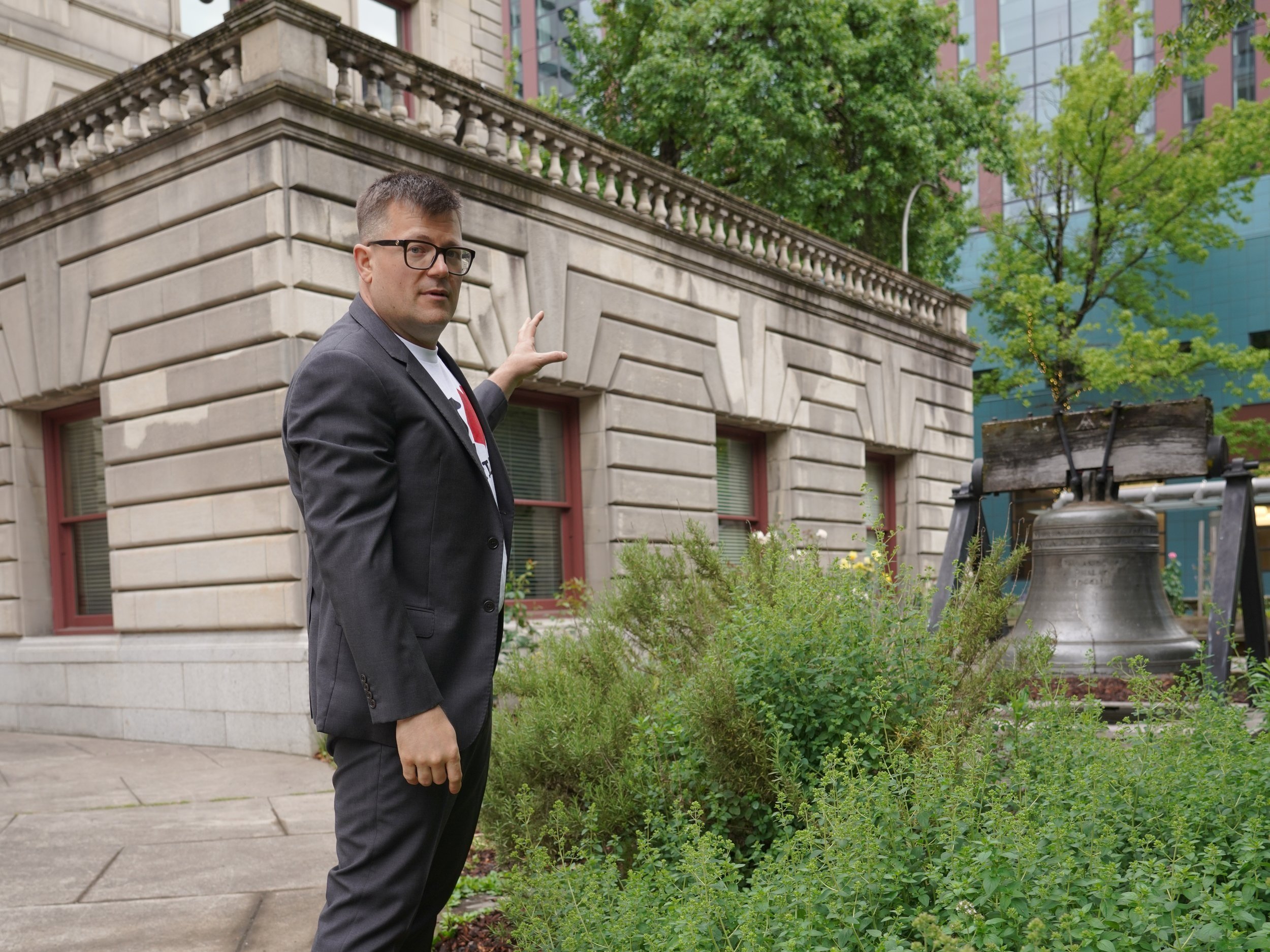 A man in a black suit and glasses gesture towards a stone building with a decorative bridge at the top, outdoor greenery, and a modern glass building in the background.