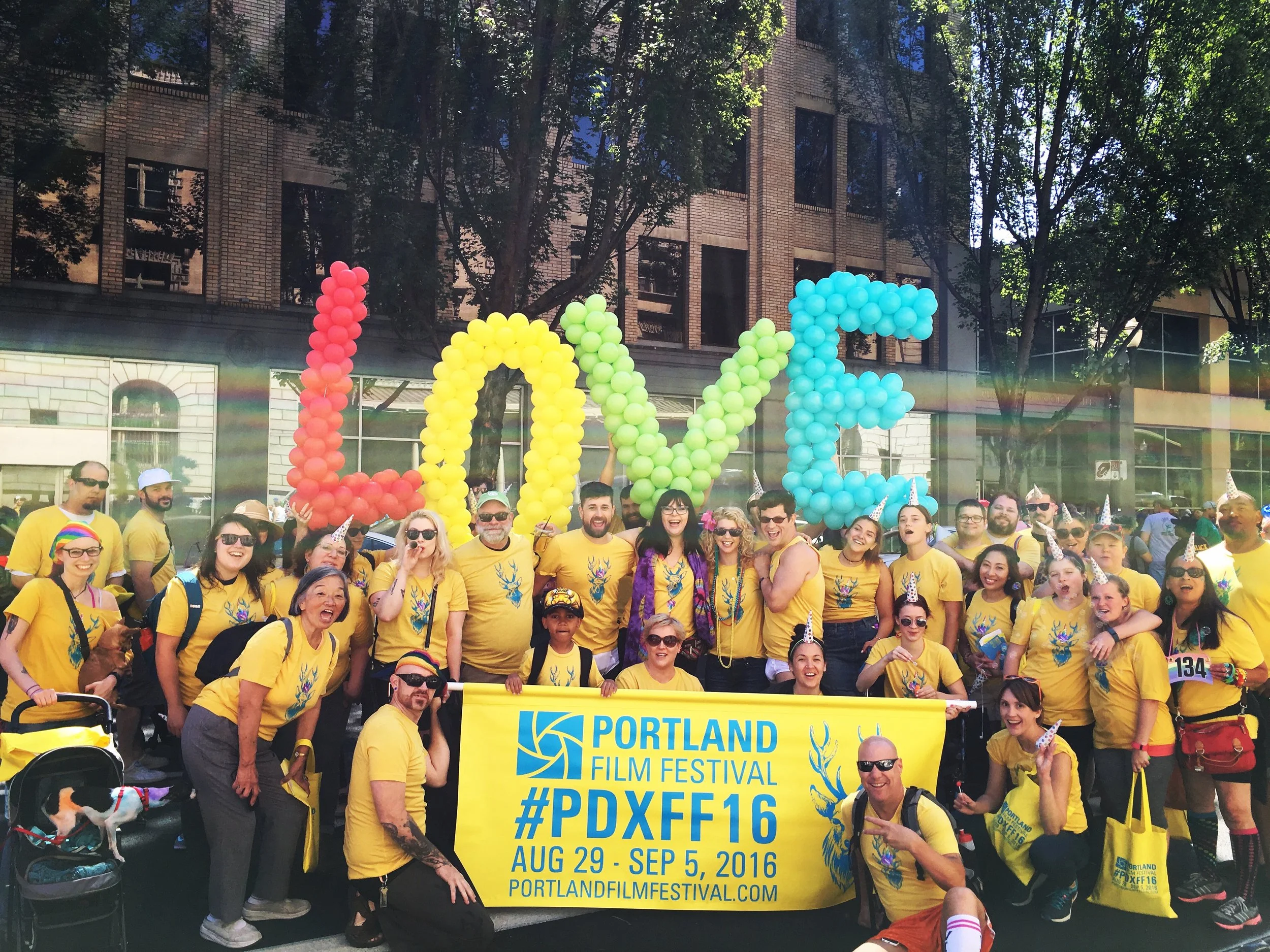 A large group of people wearing yellow shirts and party hats gather outdoors in front of a brick building with trees. They are holding a yellow banner with blue text promoting the Portland Gay and Lesbian Film Festival, August 29-September 5, 2016.