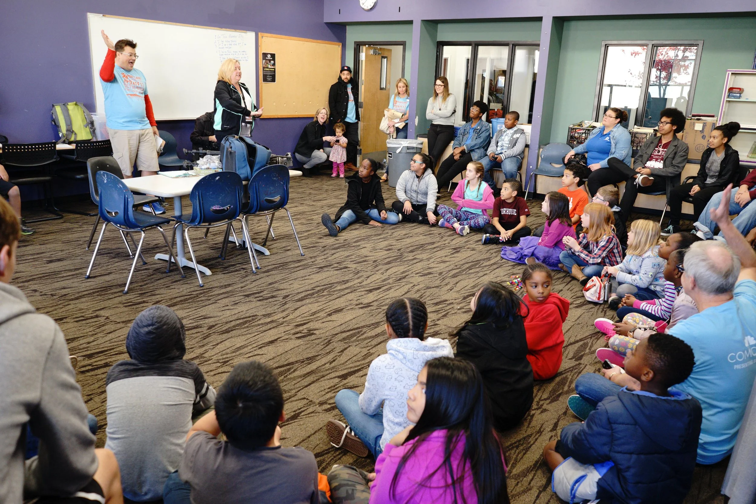 A group of children and adults gathered in a room, with some sitting on the floor and others seated on chairs, listening to a person speaking at the front. The room has colorful walls, a whiteboard, and large windows.