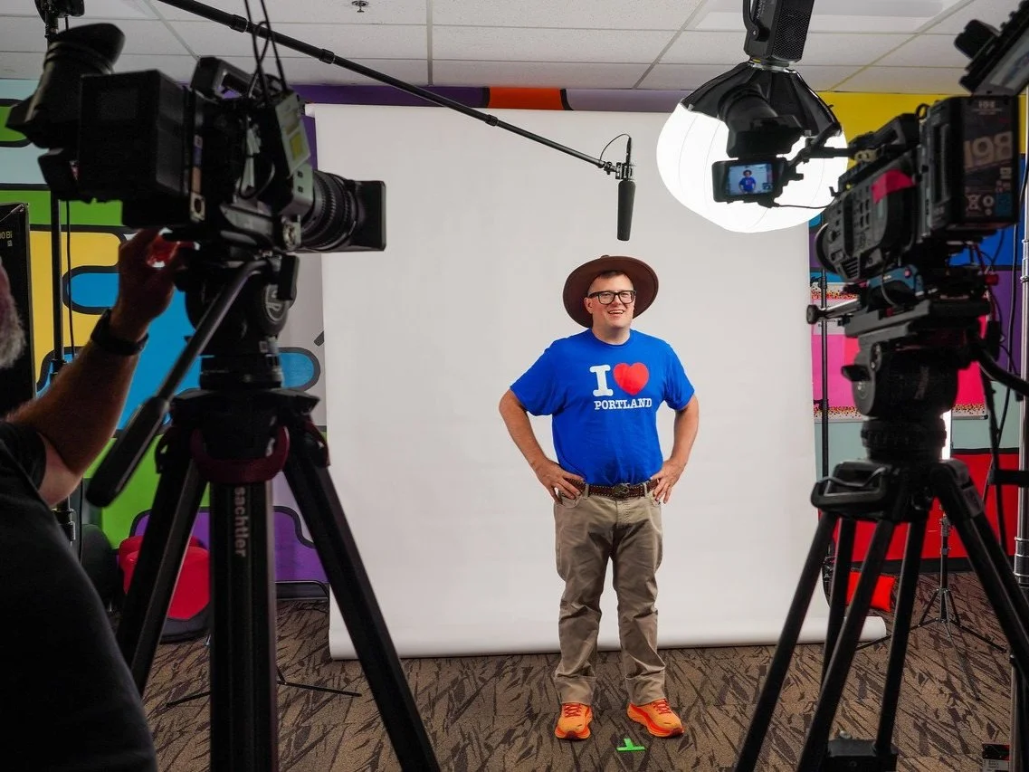 Man wearing a brown hat and glasses standing with hands on hips in front of a white backdrop, wearing a blue shirt that says 'I love Portland', beige pants, and orange shoes, during a video or photo shoot with cameras and lighting equipment.
