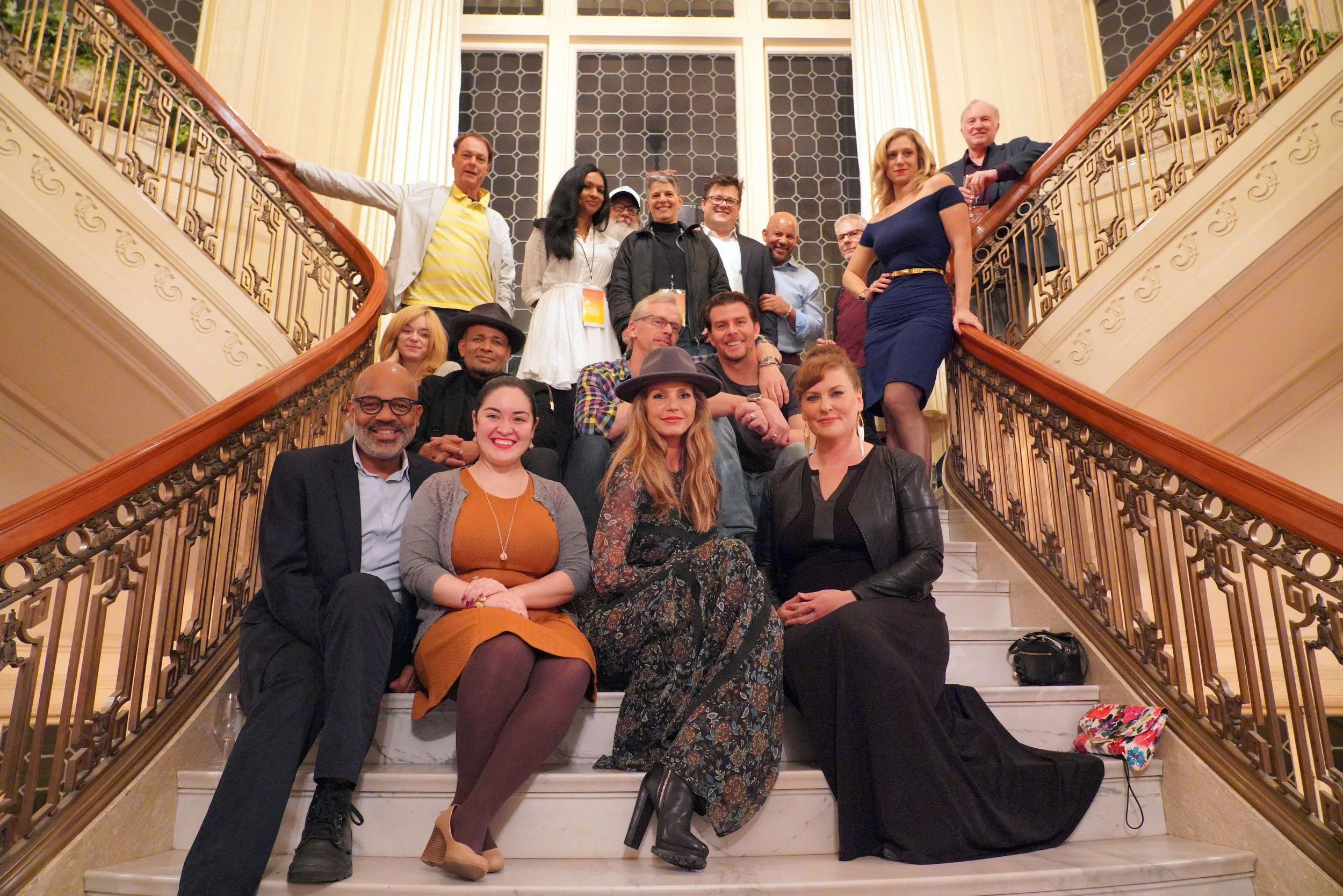 A group of 17 diverse people gathered on a staircase in a grand indoor setting with ornate railings and large windows, posing for a photo during an event.
