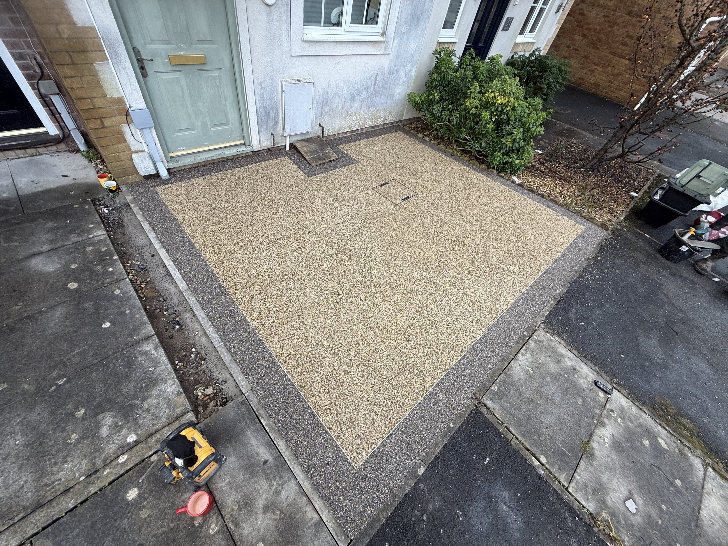 View of a small front yard with a new decked area in front of the house door, surrounded by a concrete border. The yard has a green bush next to the house, and to the right, two trash bins and a few bags are placed on the sidewalk. The house has a gr