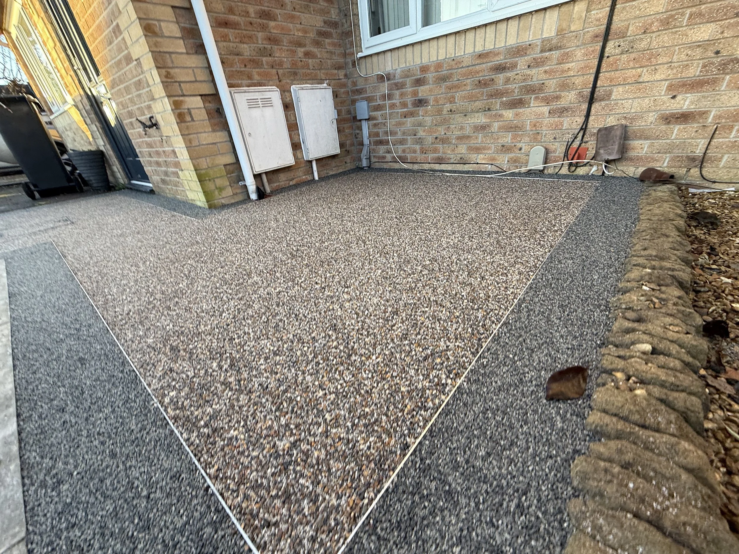 Newly paved gravel patio area next to a brick house with utility boxes and wires, bordered by a row of stones.