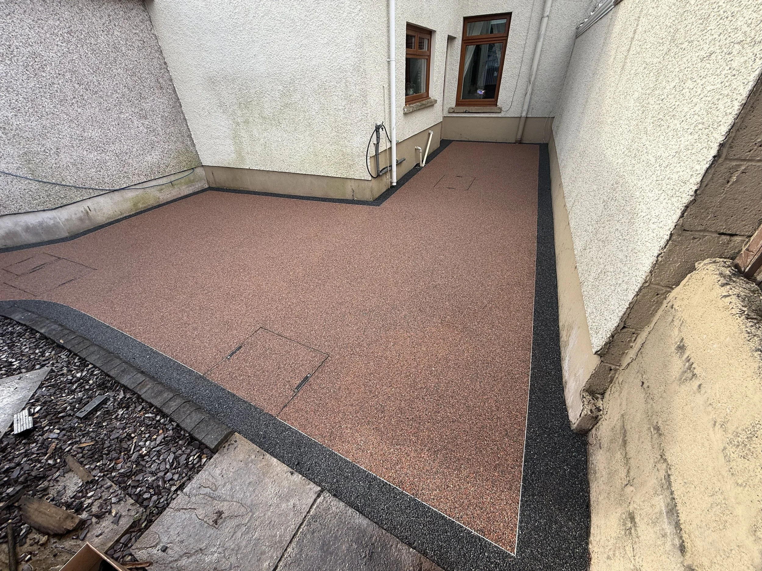Empty small balcony with reddish-brown textured flooring, black border, and white stucco walls, with two small windows and drain covers.