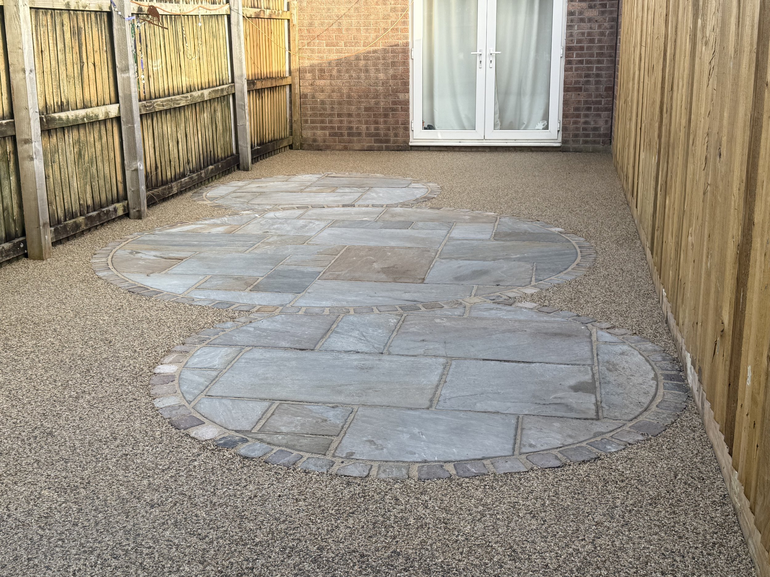 Backyard with newly paved stone patio and surrounding gravel ground, enclosed by wooden fences and a brick wall with a patio door.