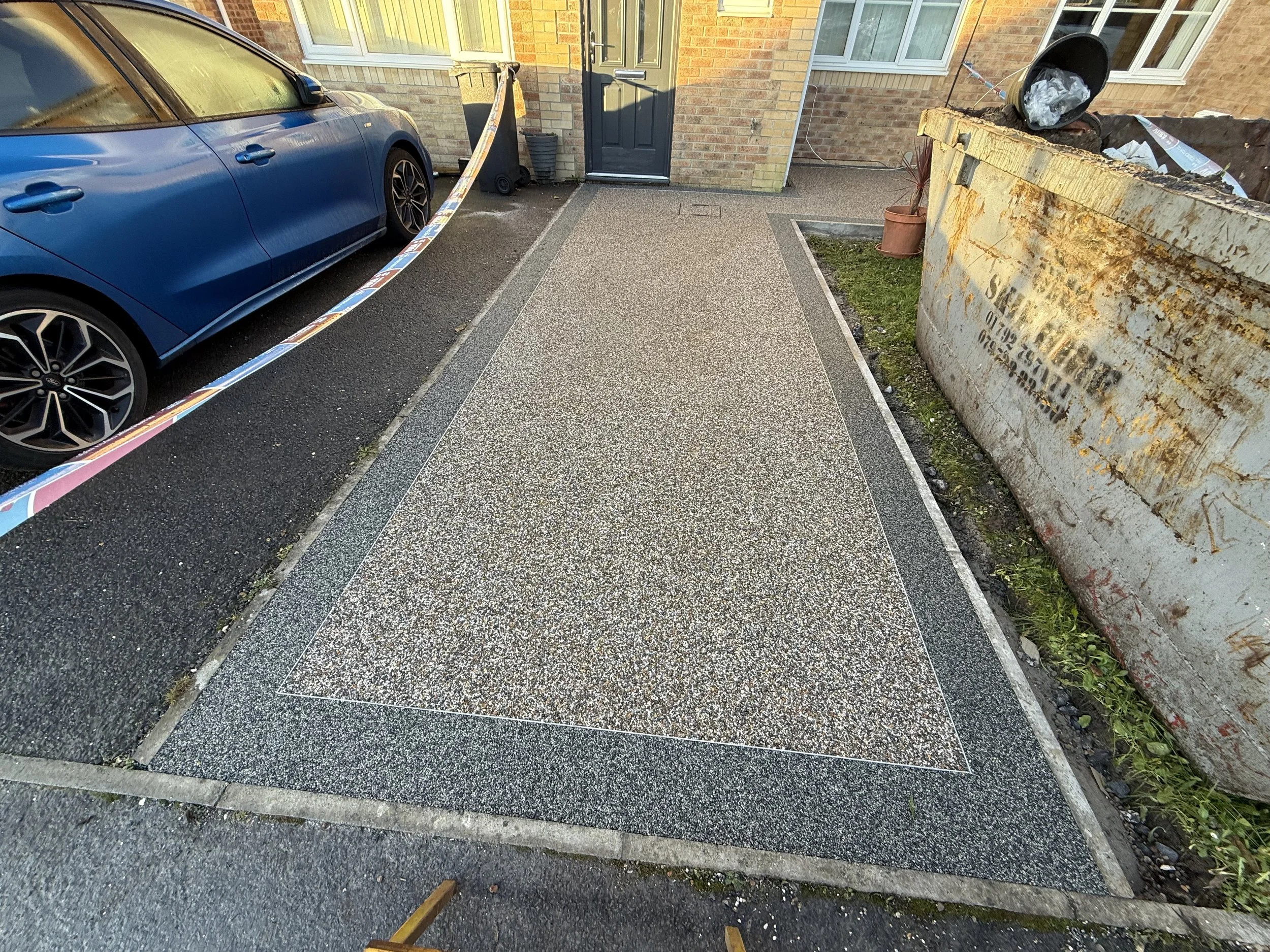 View of a newly paved or renovated concrete driveway outside a house, with a blue car parked on the left, a black security camera on the house wall, and a rusty old service dumpster on the right. The driveway has a central rectangular design, bordere