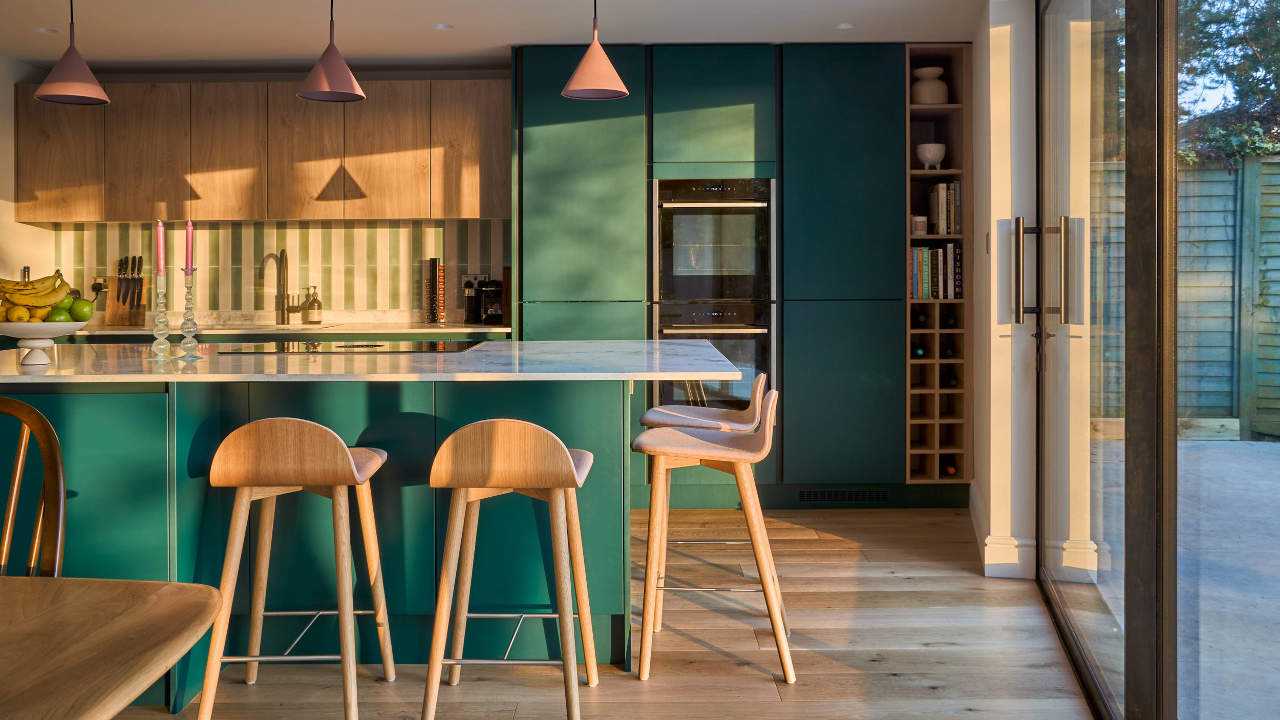 Modern kitchen with green cabinets, wooden accents, marble countertop, and sunlight coming through glass sliding doors.