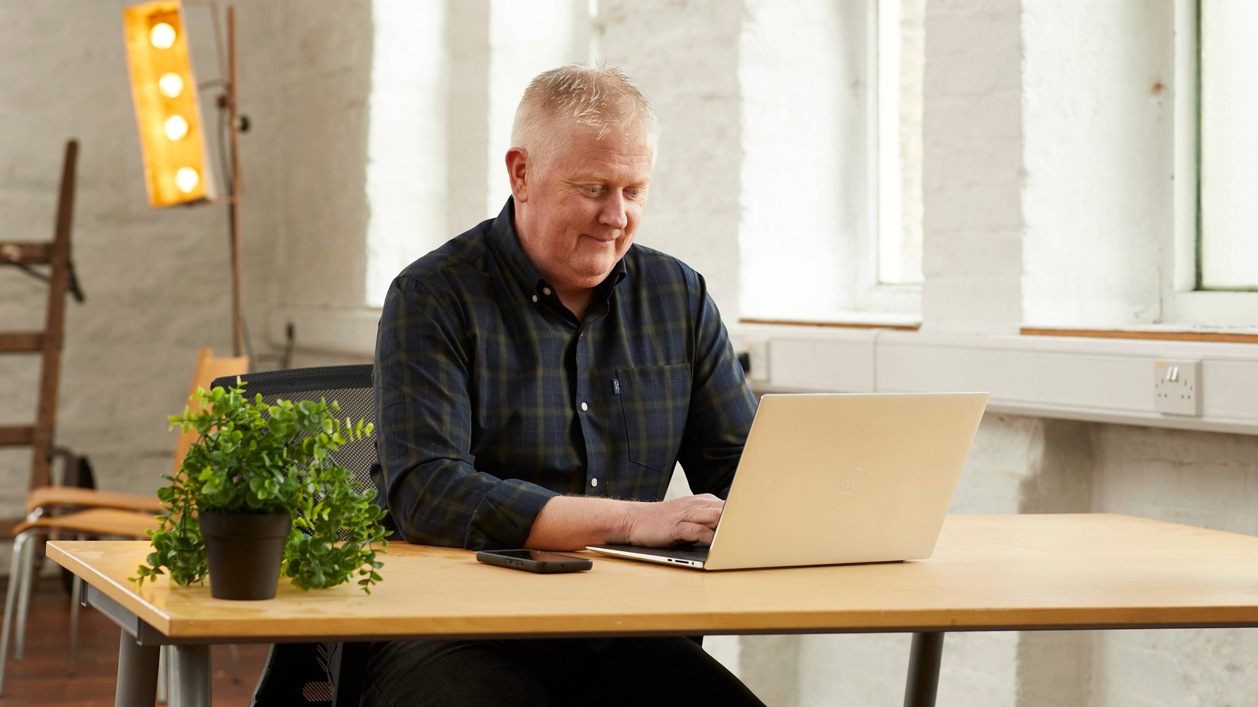 A man working on a laptop at a wooden desk in a bright, industrial-style room with white brick walls and large windows.