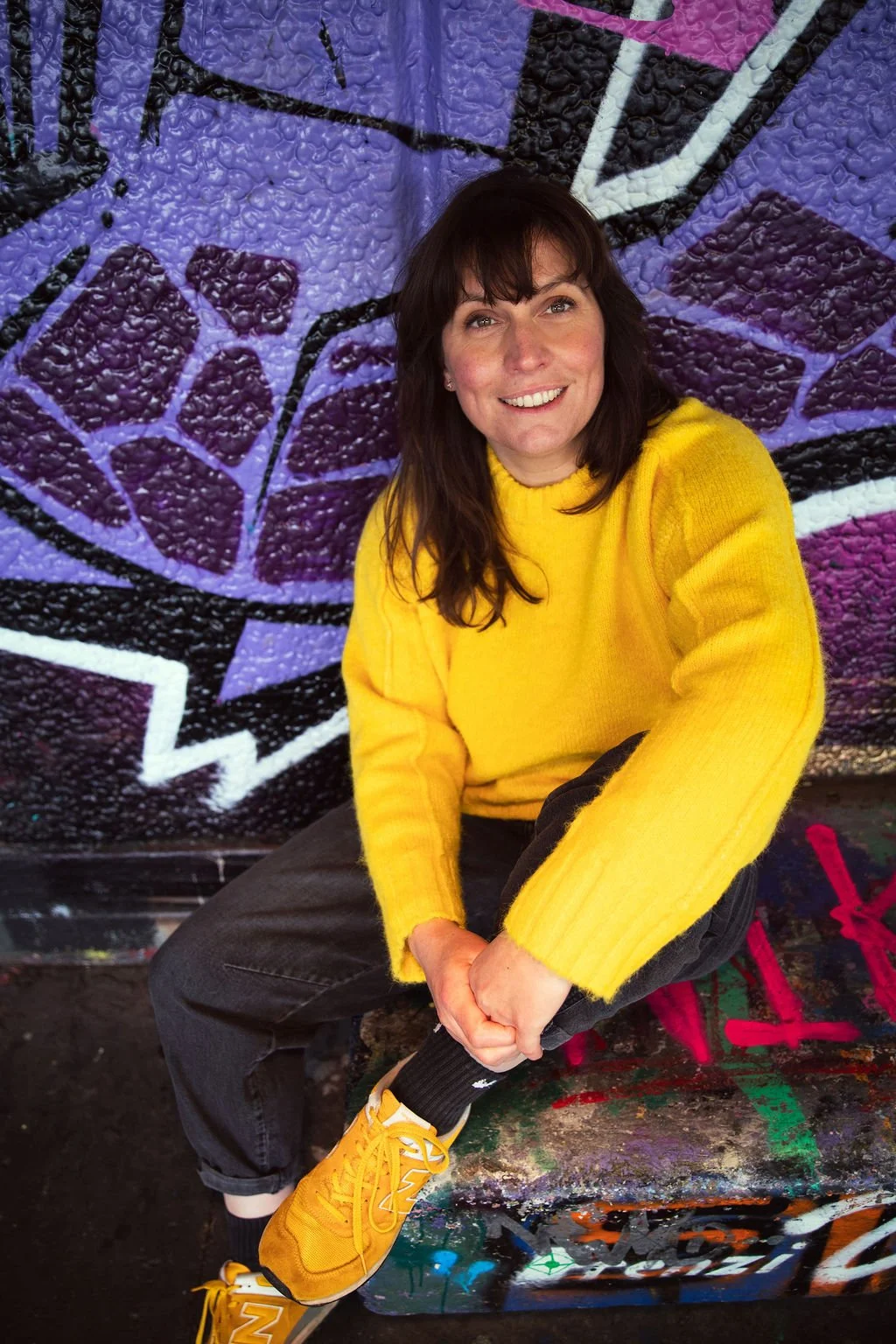 A woman wearing a yellow sweater and yellow sneakers sitting in front of a colorful graffiti wall and smiling.