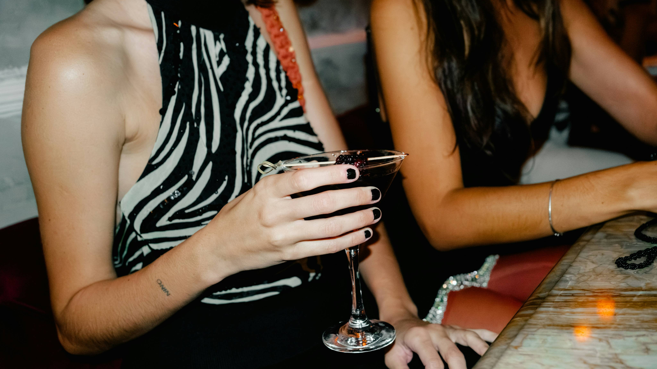 A woman holding a martini glass with dark berries at a bar or restaurant, with another woman sitting beside her. Both are dressed in black, with one wearing a zebra-print top.