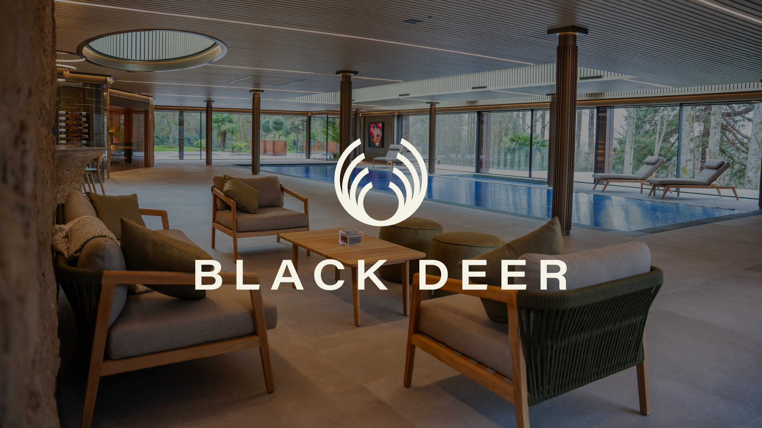 Interior view of a luxury indoor pool area at Black Deer with brown chairs, a wooden table, lounge chairs by the pool, and large glass windows showing outdoor greenery.