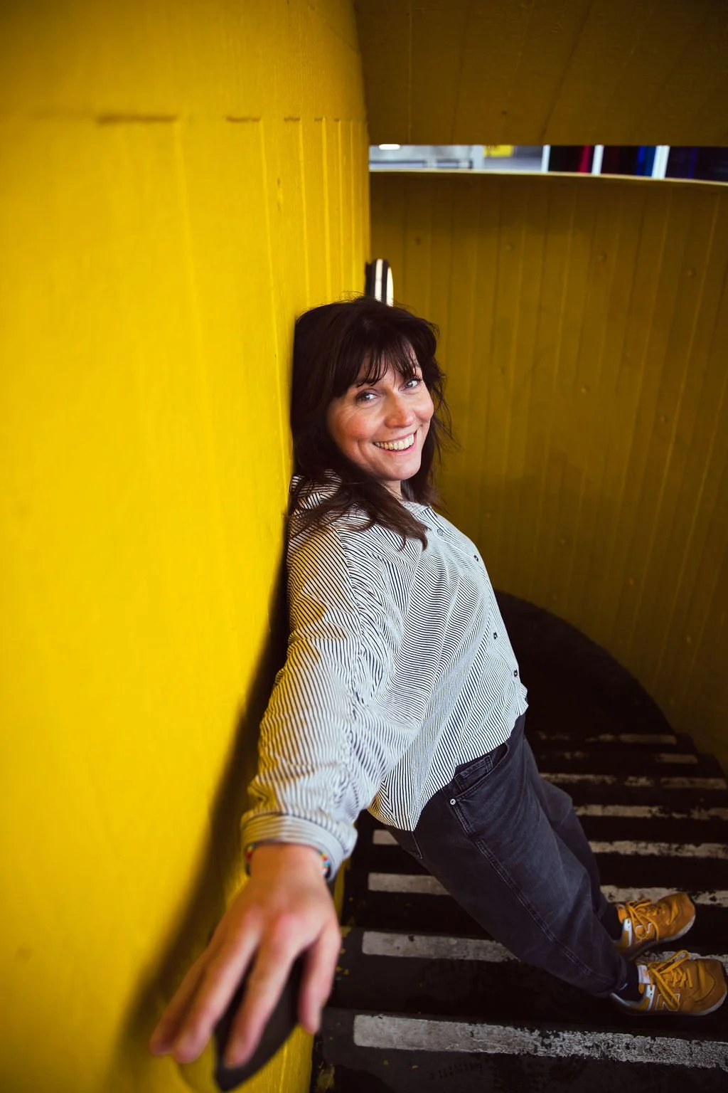 A woman with dark hair and a striped shirt smiling and leaning against a yellow wall in a stairwell.