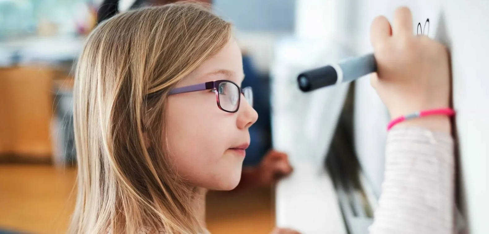 A young girl with glasses writing on a whiteboard with a black marker.