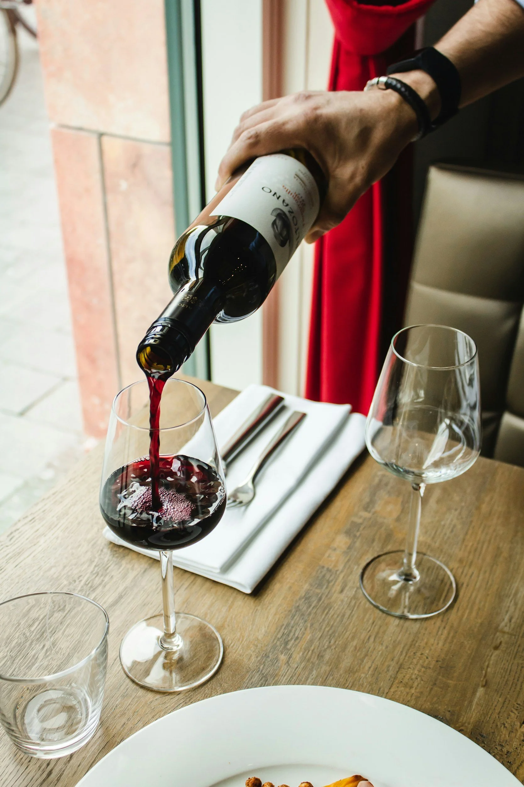 Une personne verse du vin rouge dans un verre à vin sur une table en bois, avec une assiette, un verre d'eau vide, des couverts et un verre à vin vide à côté.