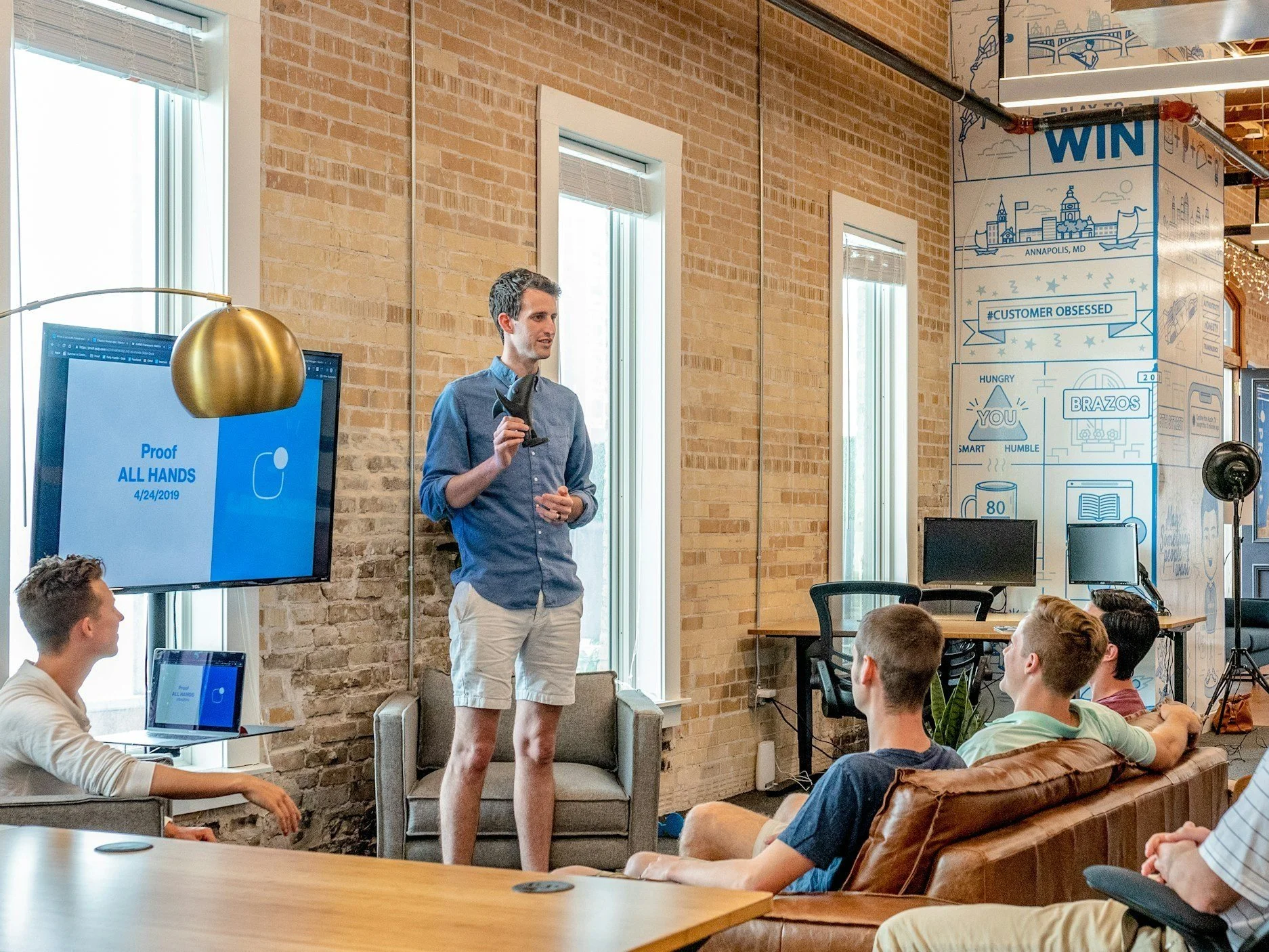 Une présentation sur la lutte contre la COVID-19, affichant 'Proof ALL HANDS 4/24/2019', dans un espace de travail moderne avec un mur en briques, un écran, un tableau interactif et des personnes assises en train d'écouter.