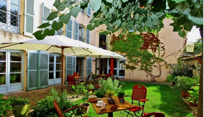 Une terrasse de café ou restaurant avec des tables, des chaises en bois peint en rouge et des parasols blancs, entourée de verdure et de plantes grimpantes, dans un environnement charmant et paisible.