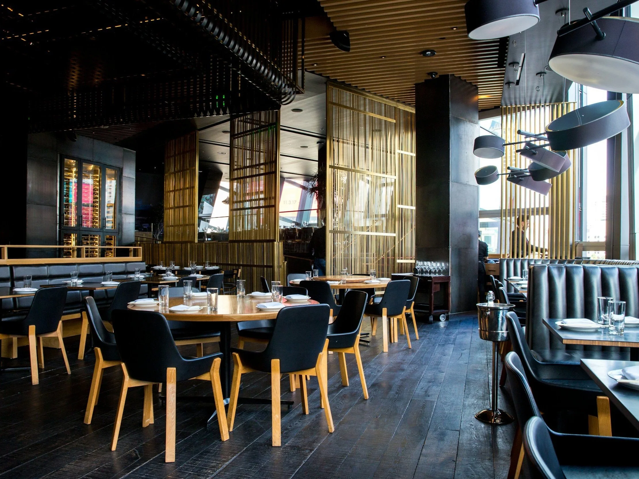 Intérieur d'un restaurant moderne avec des tables en bois, des chaises noires et un décor en bois doré, éclairage suspendu et grandes fenêtres naturelles.