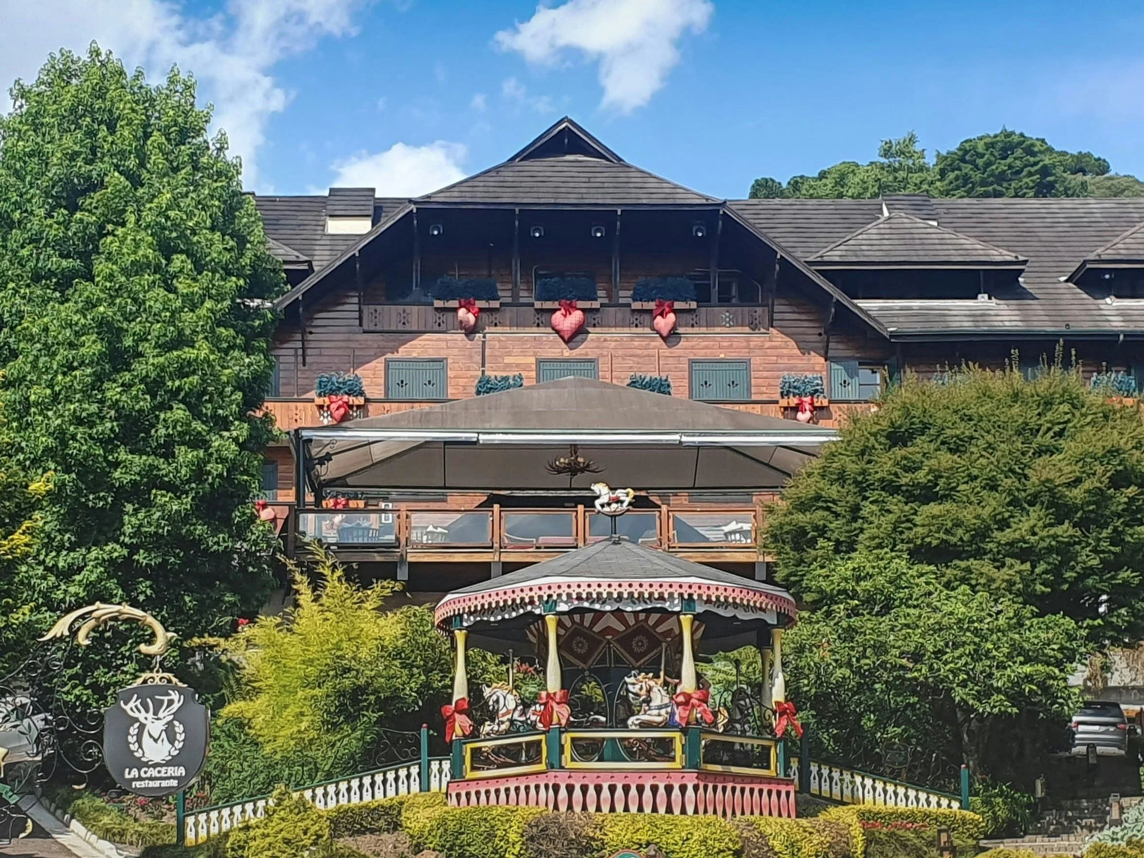 Un bâtiment en bois avec un balcon et des décorations de cœurs suspendus, un manège de chevaux en bois décoré de rubans rouges, entouré de végétation verte.