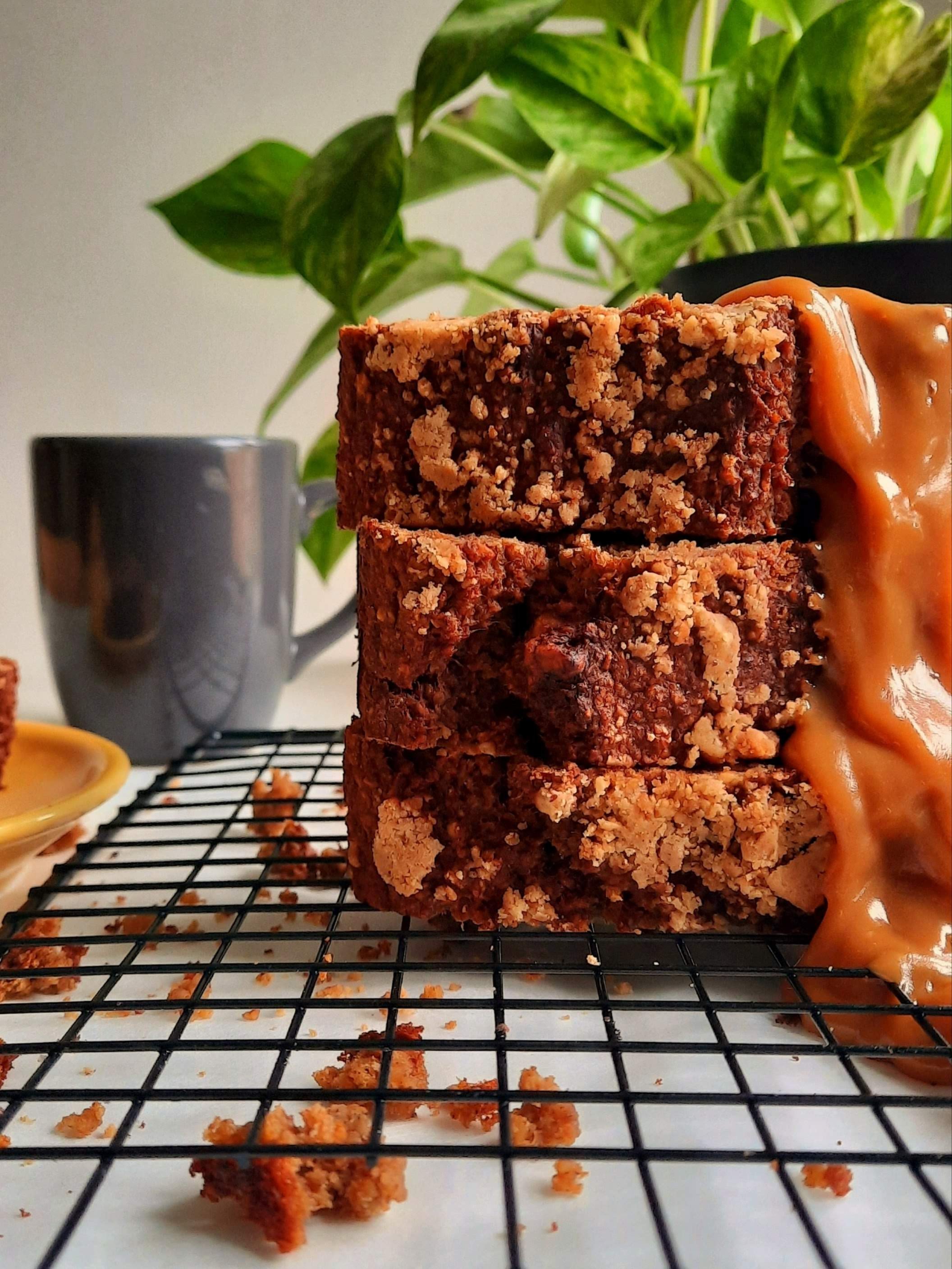 Gâteaux au chocolat avec un glaçage caramélisé, placé sur une grille, avec des miettes autour et une tasse gris foncé derrière, une plante verte en arrière-plan.