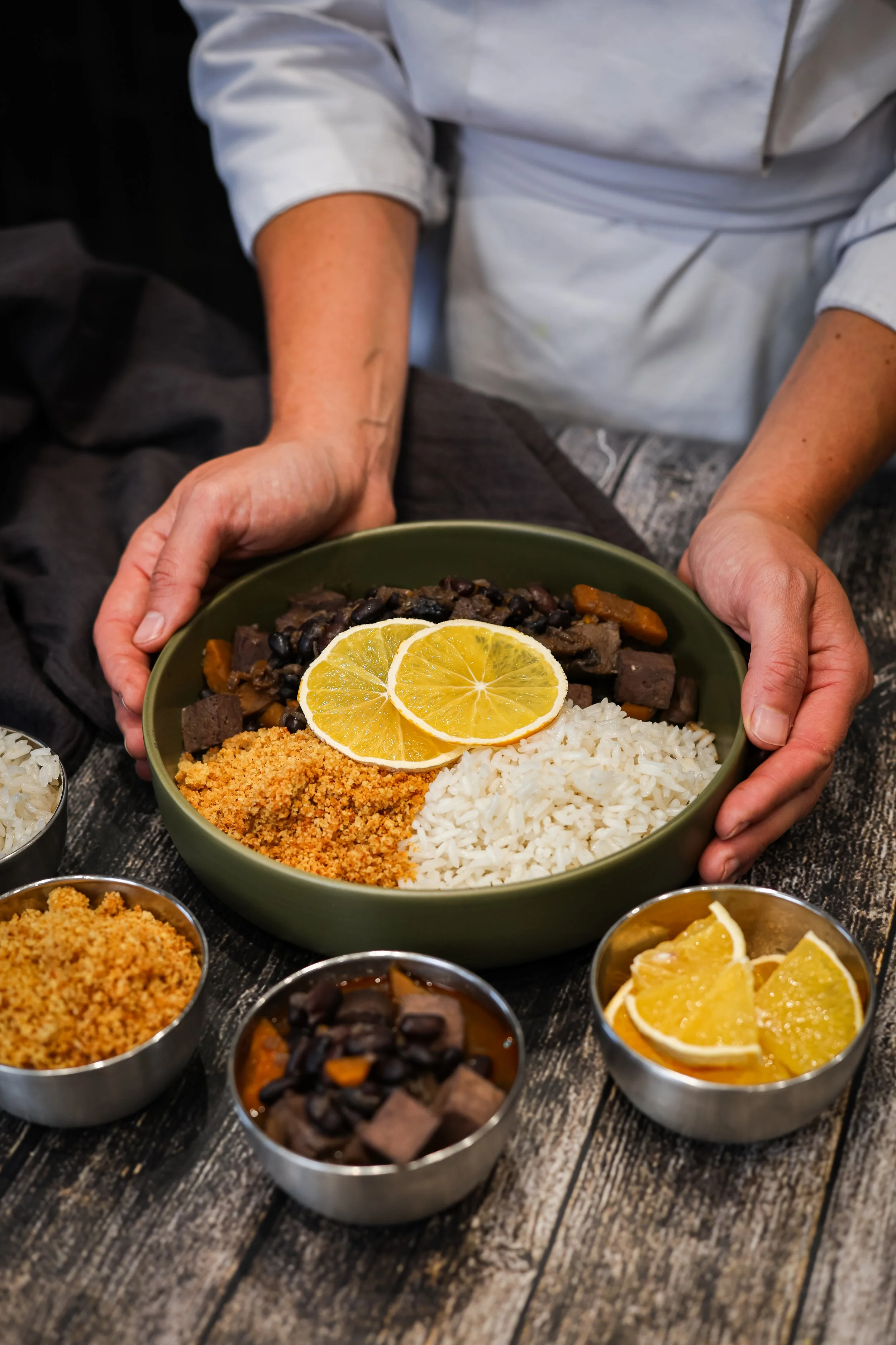 Une personne présente un plat de riz blanc, bananes en tranches, haricots noirs, viande et autres accompagnements sur une table en bois.
