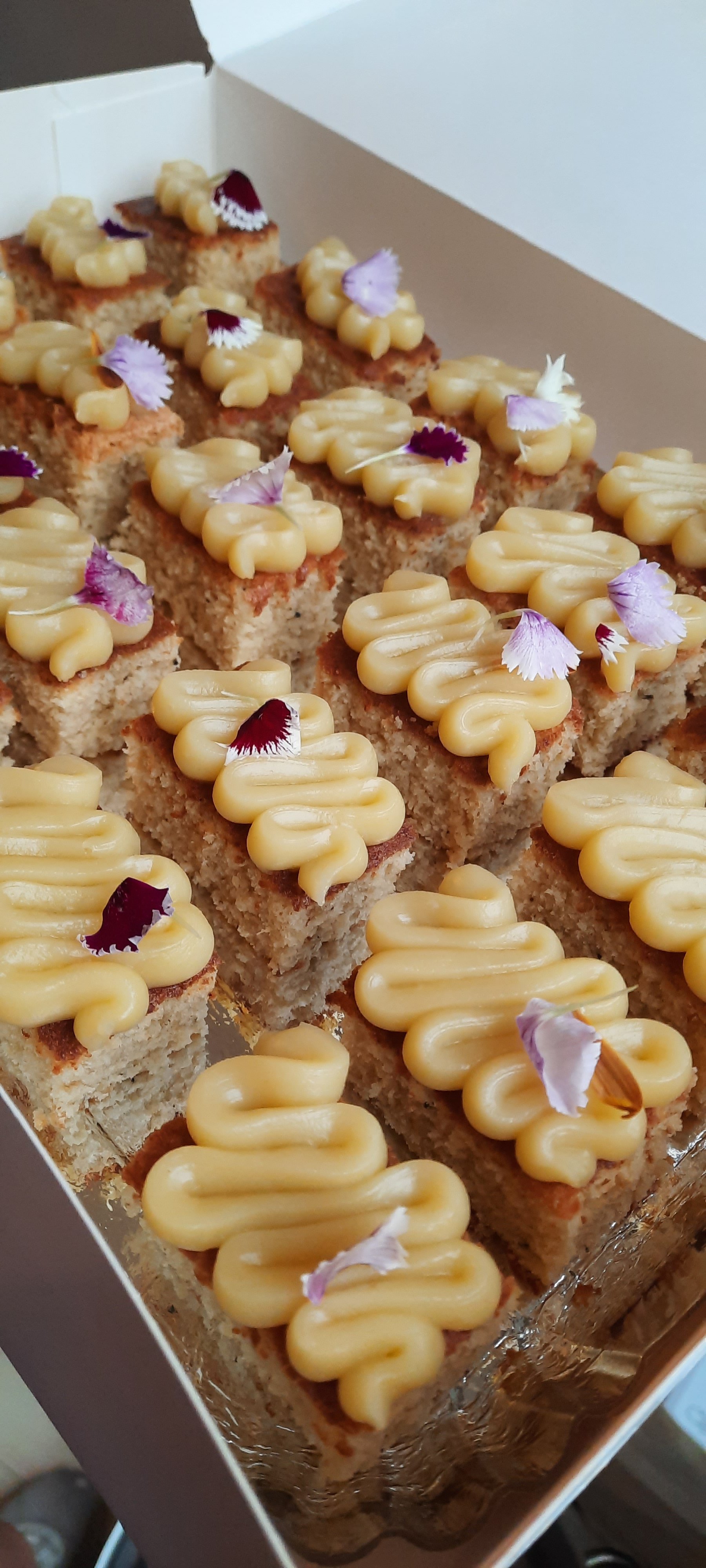 Gâteau en forme de carrés, décoré avec de la crème pâtissière et des fleurs comestibles violettes, en boîte en carton.