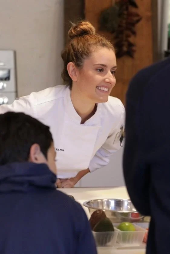 Carla Viana en uniforme de cuisine souriante, discutant avec deux personnes, dans une cuisine moderne.