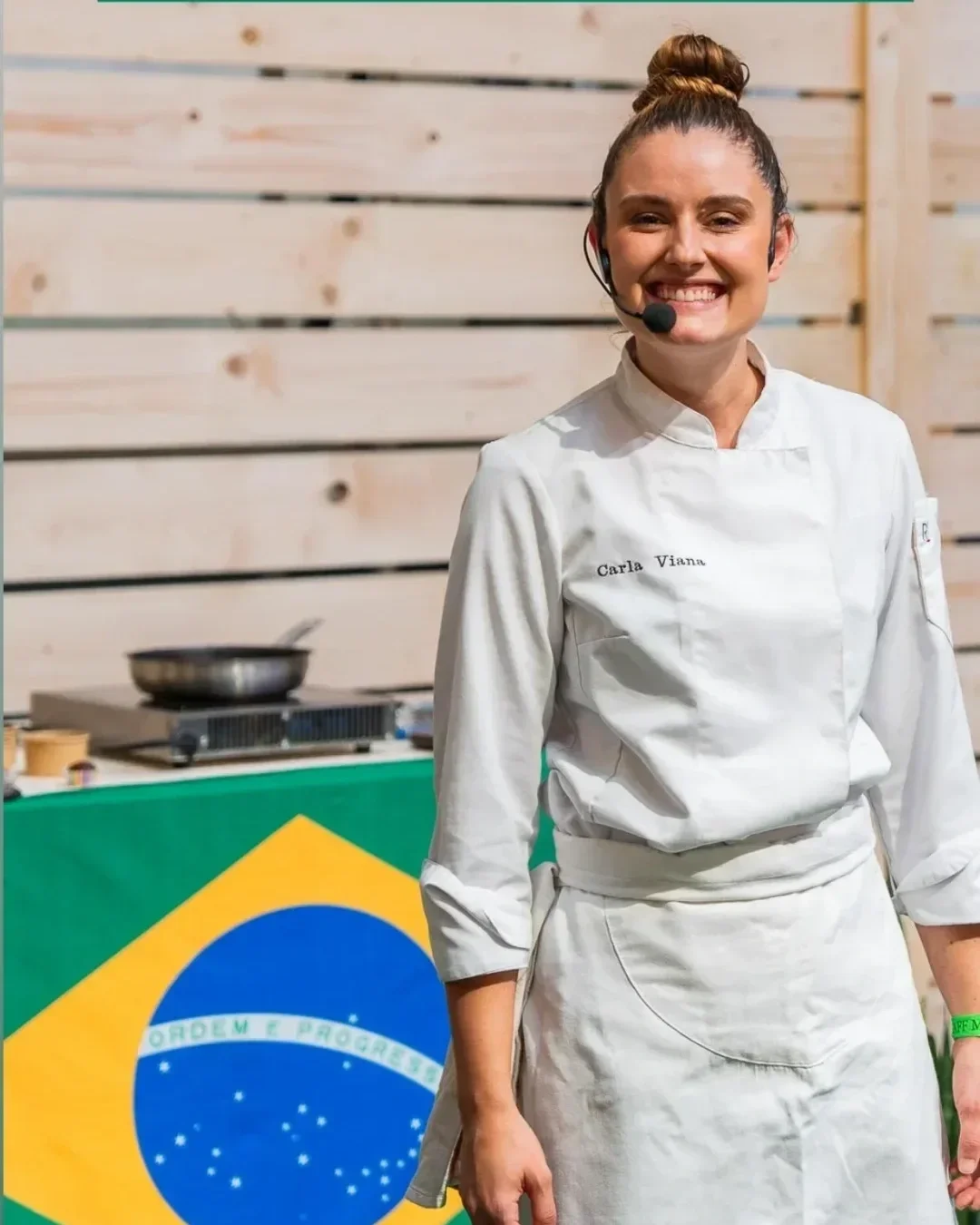 Carla Viana souriante en costume de chef, portant un micro-casque, se tient devant un drapeau du Brésil.