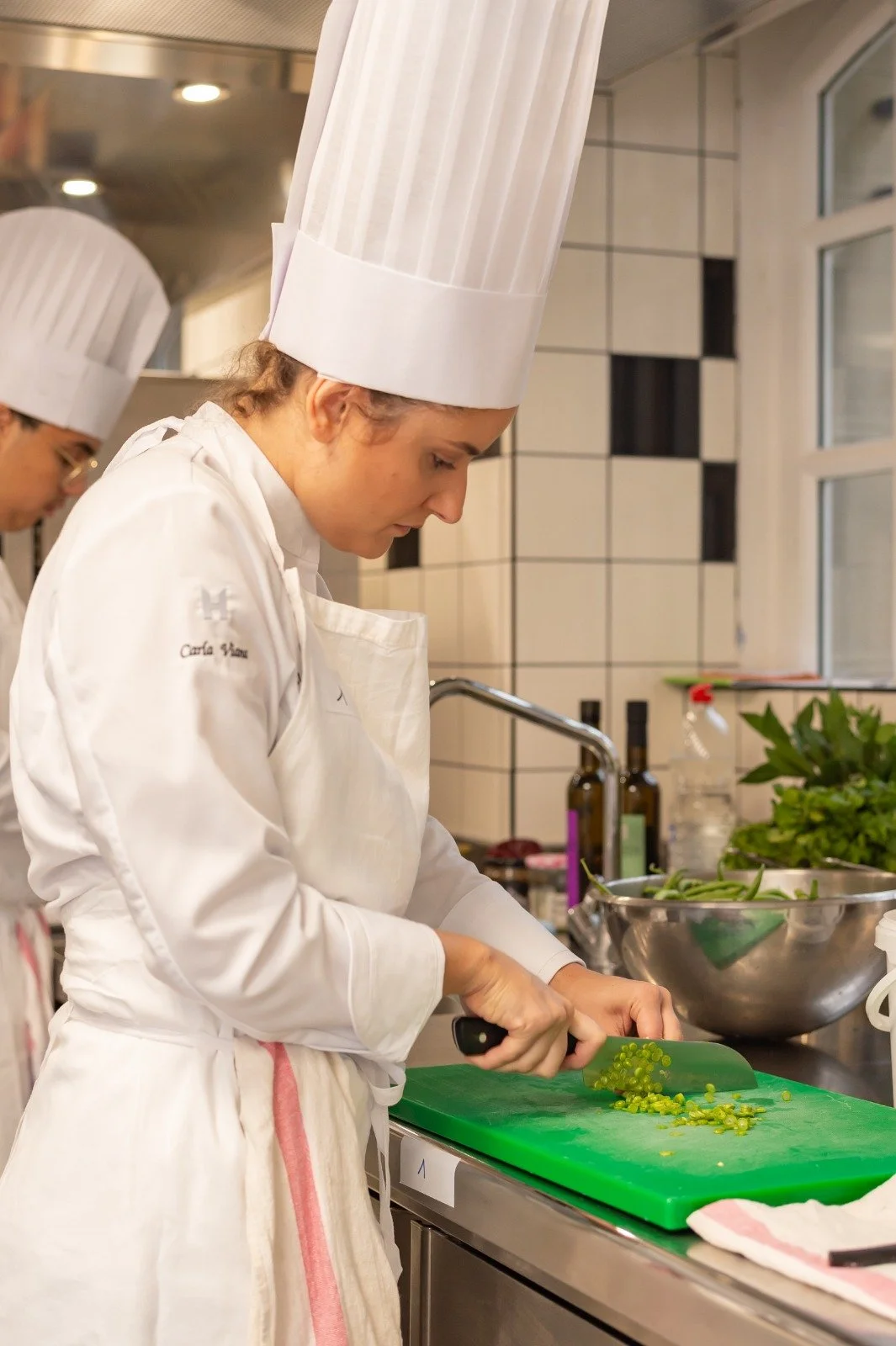 Carla Viana en tenue blanche coupe des légumes verts sur une planche à découper dans une cuisine professionnelle.