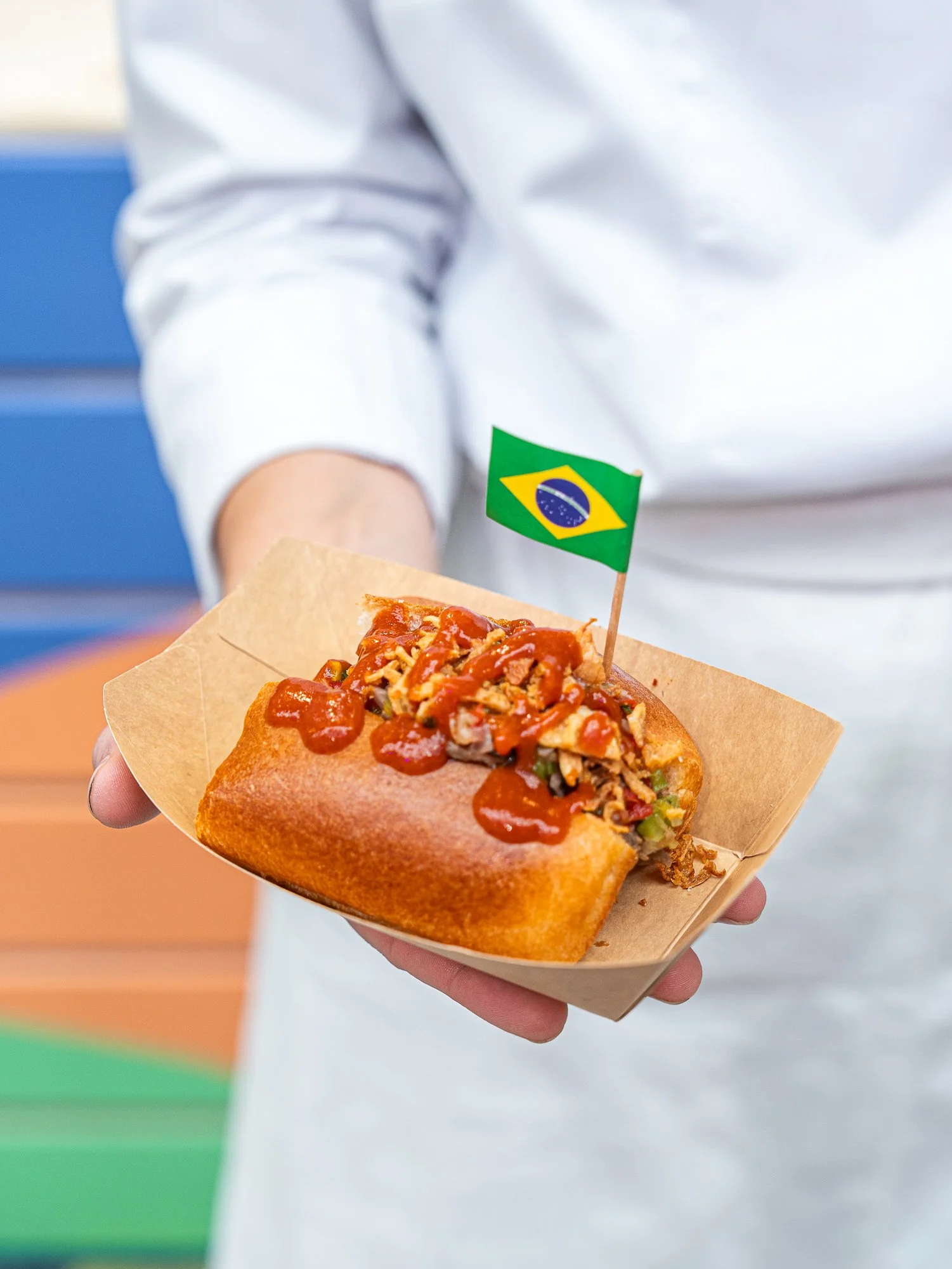 Une personne tient un pão de queijo brésilien garni de sauce, avec un drapeau du Brésil planté dessus, dans un support en papier.